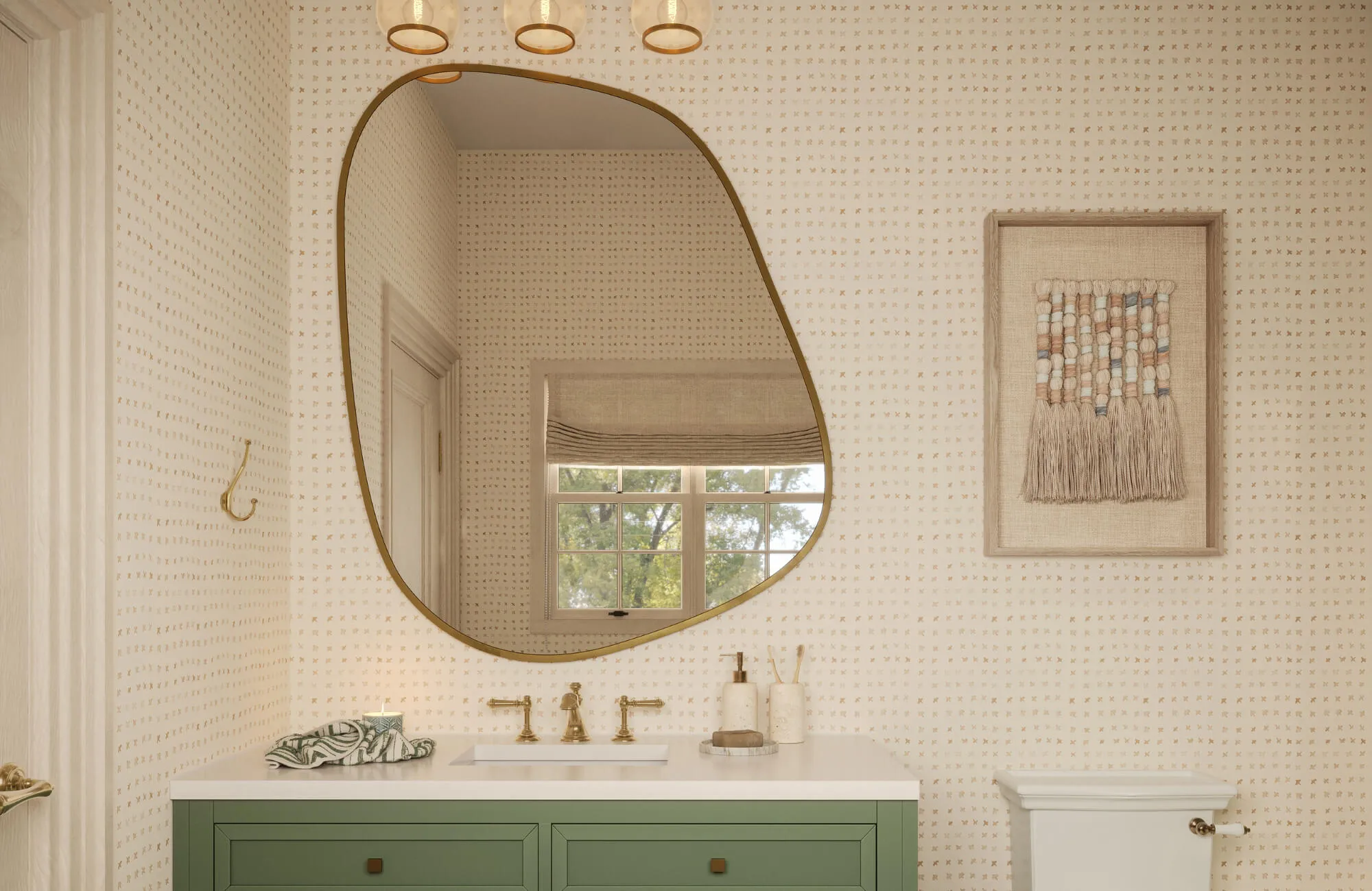 Modern bathroom with asymmetrical brass mirror, sage vanity, woven wall art, and patterned wallpaper accents throughout the room.