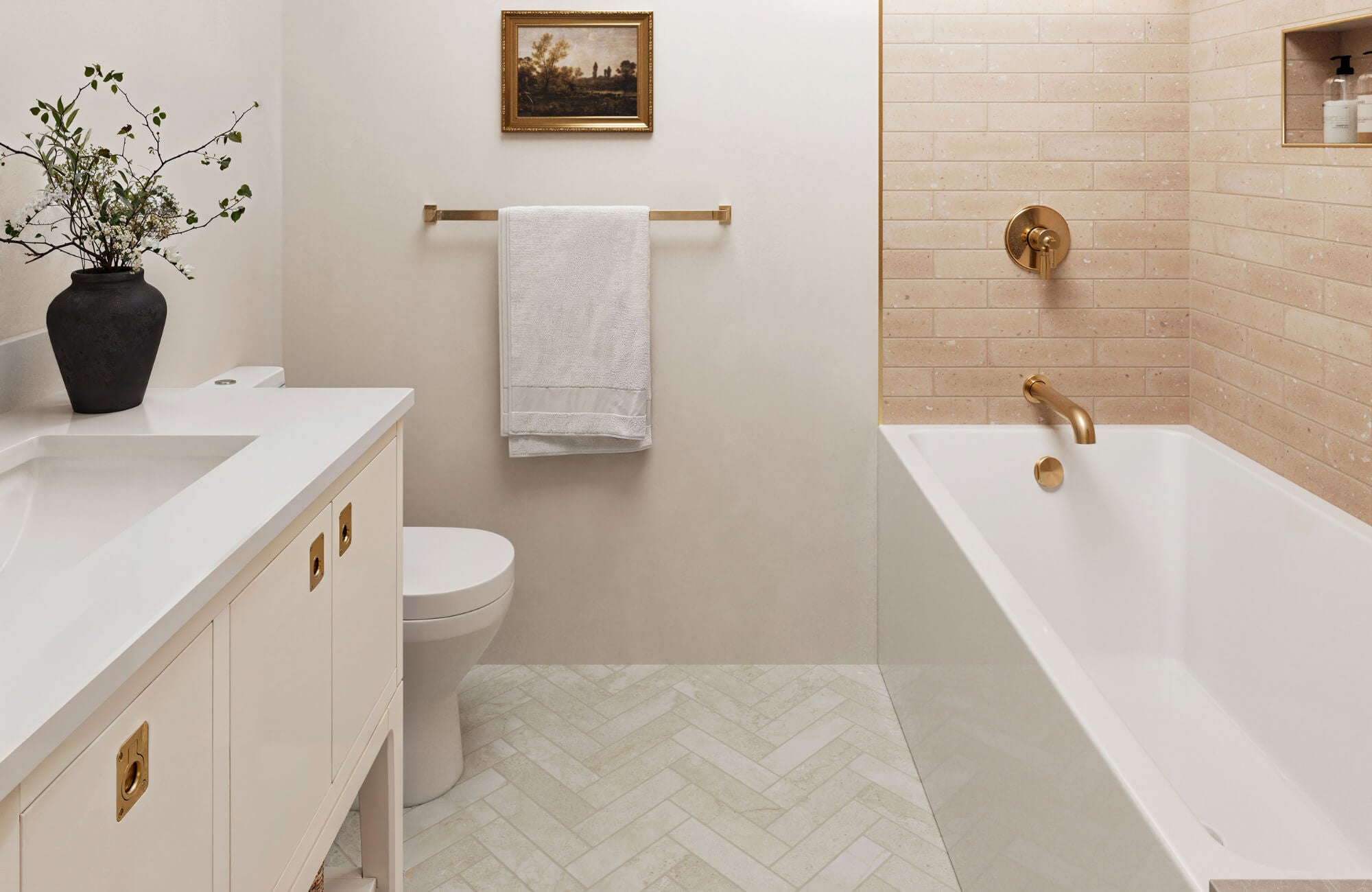 Elegant bathroom with herringbone ceramic tile flooring, gold fixtures, a white vanity, and a bathtub with warm beige subway tile walls.