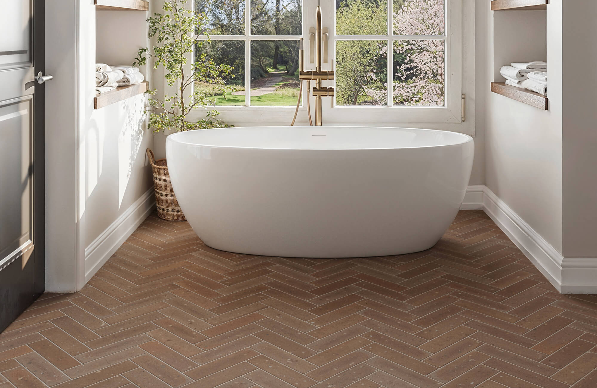 Bright bathroom with a freestanding tub, warm brown herringbone ceramic tile flooring, and large windows overlooking a garden view.