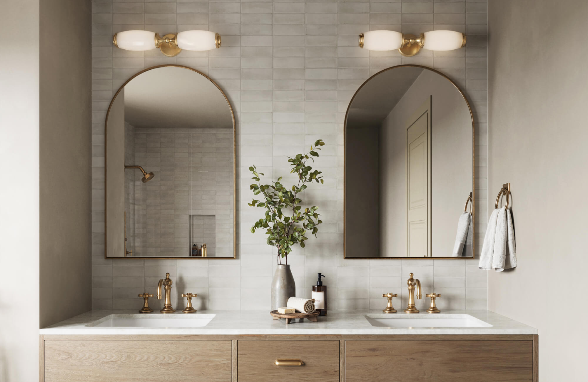 Bathroom featuring dual sinks, warm brass hardware, arched mirrors, and wall-mounted lights on a neutral tiled backsplash.