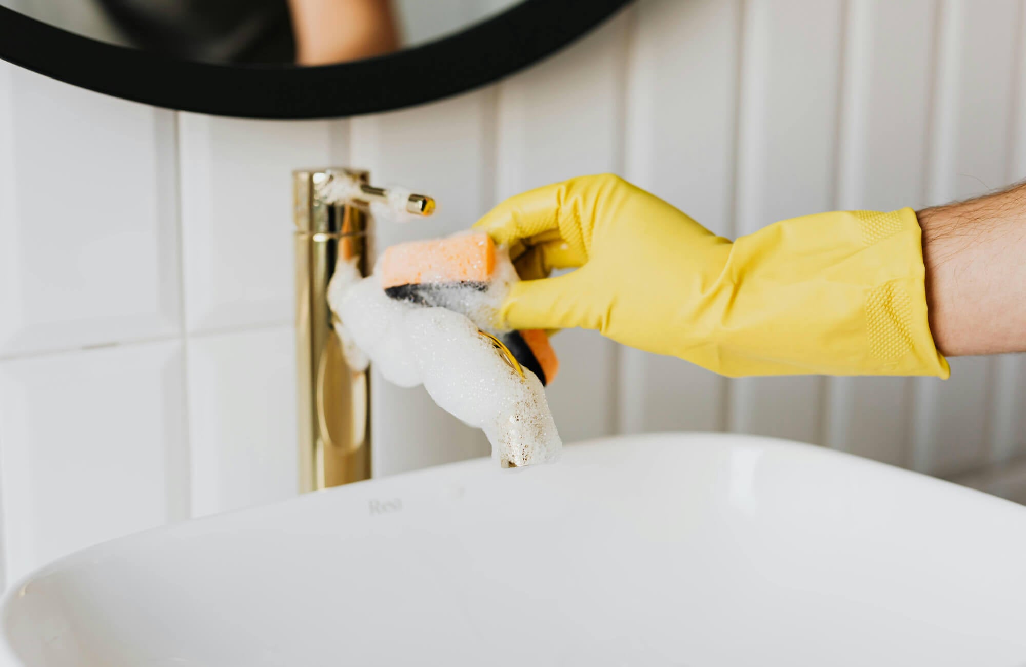 Hand in a yellow glove, scrubbing a gold bathroom faucet with a sponge and soap suds.