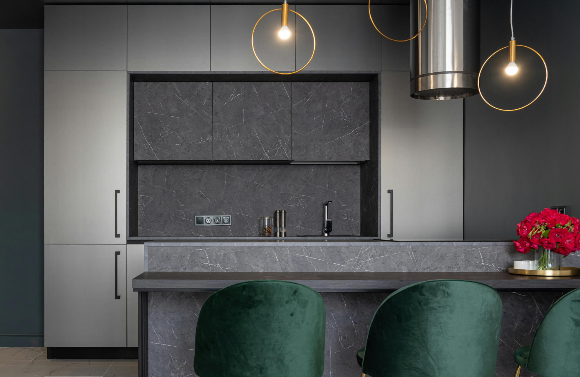 Sleek modern kitchen highlighted by minimalist ring-style kitchen pendant lighting above a gray stone island, complemented by matte cabinetry, dark marble surfaces, and rich green bar stools for a bold, contemporary aesthetic.