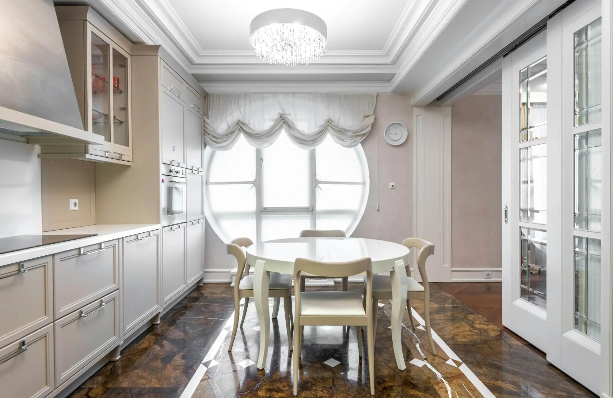 Alt text: Elegant kitchen dining area with glossy brown marble flooring, white cabinetry, a round white table with four chairs, and a large circular window dressed in soft drapery.