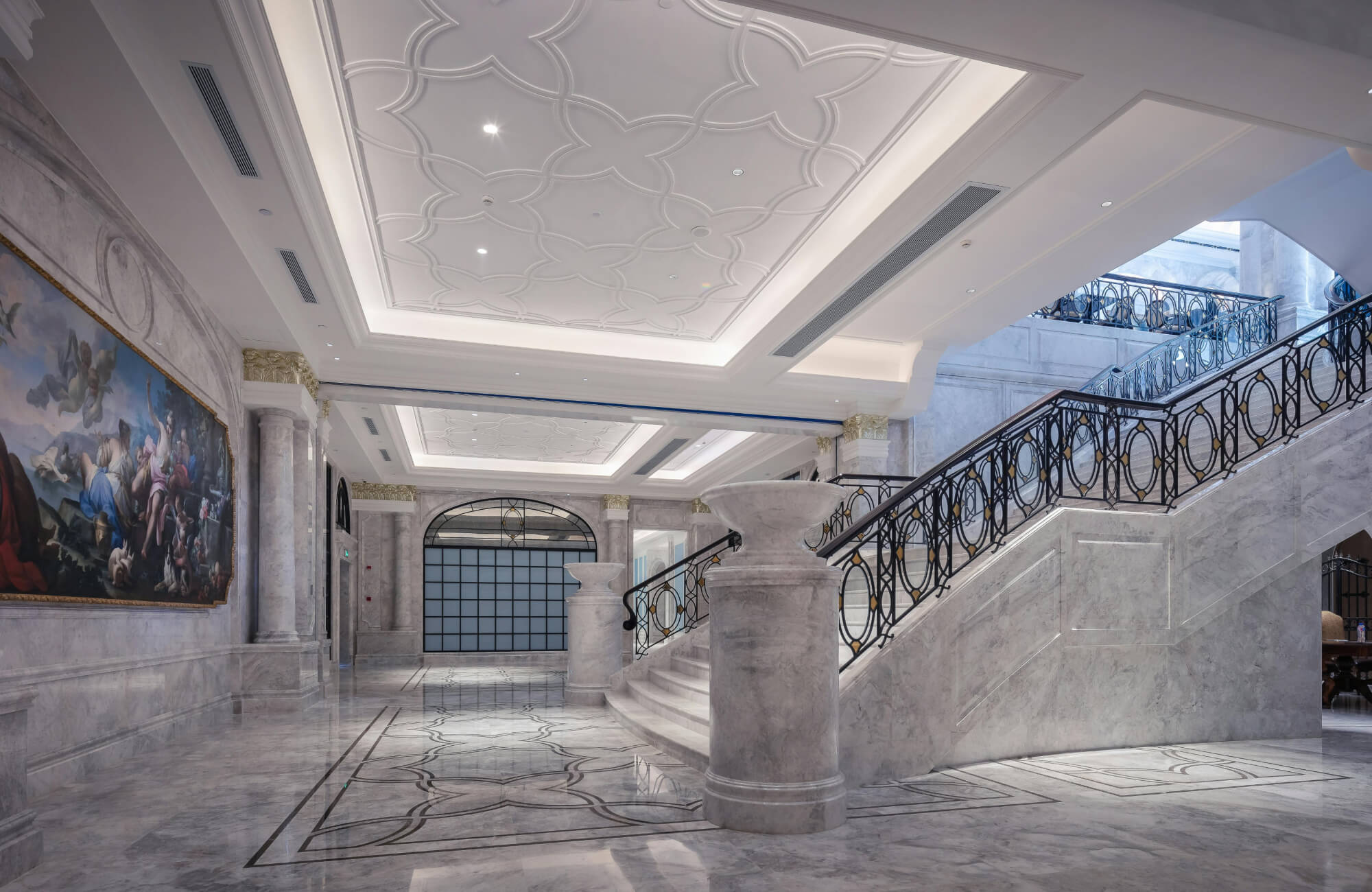 Grand lobby with luxurious white marble flooring featuring intricate black inlays, complemented by ornate columns, an elegant staircase, and a striking ceiling design.