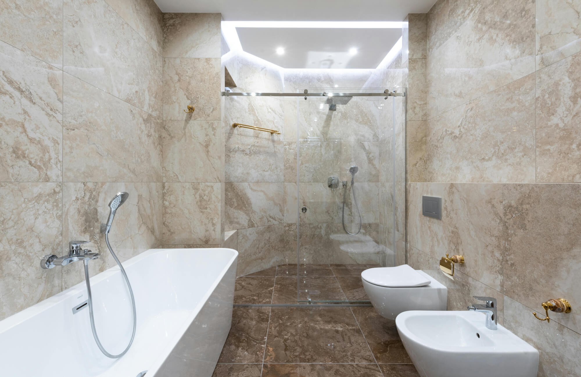 Luxurious bathroom with polished beige marble walls and contrasting brown marble flooring, featuring a glass-enclosed shower and elegant gold accents.