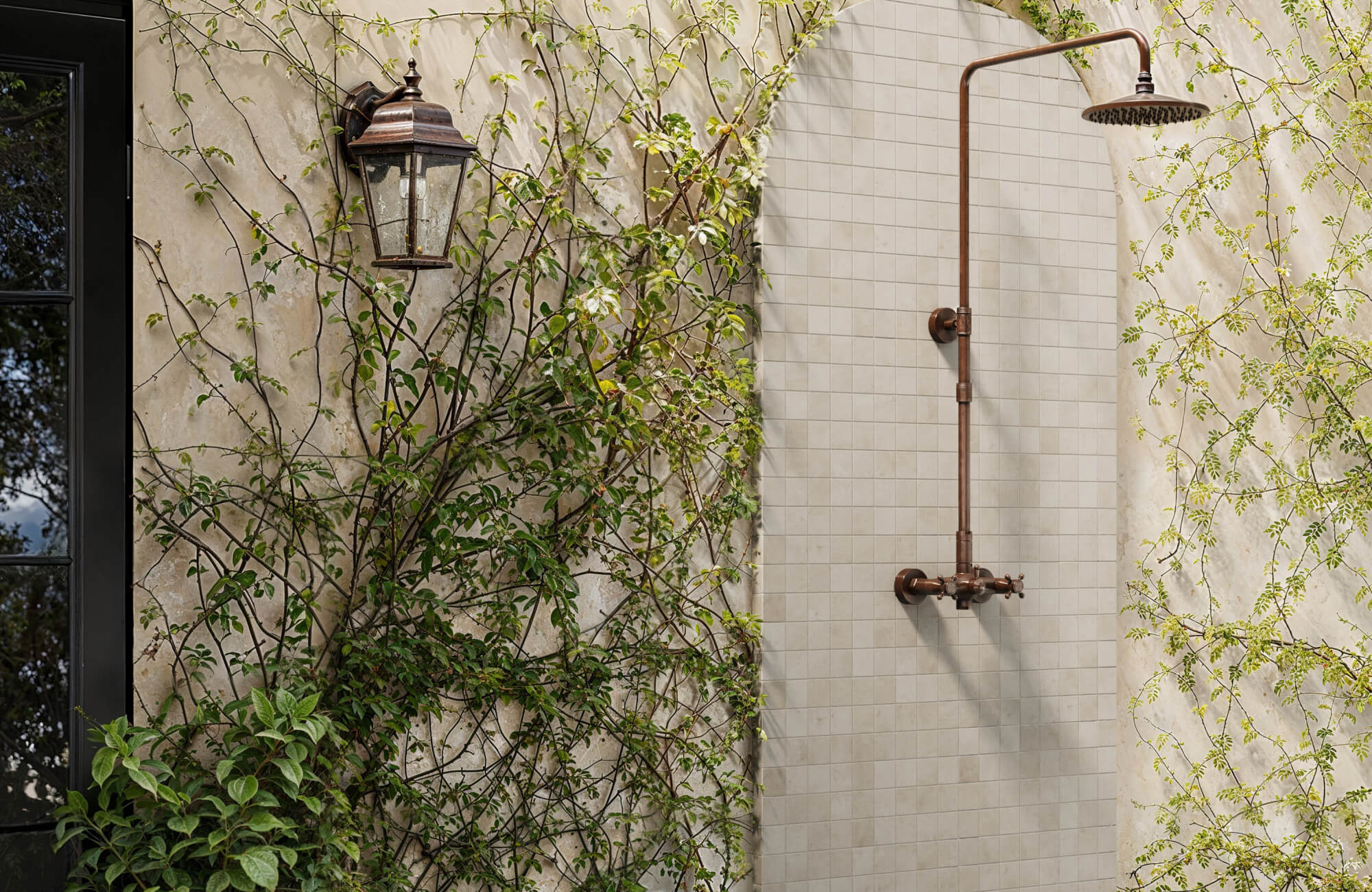 Outdoor shower framed by warm beige square mosaic tiles, set against a vine-covered stucco wall for a rustic, natural retreat.
