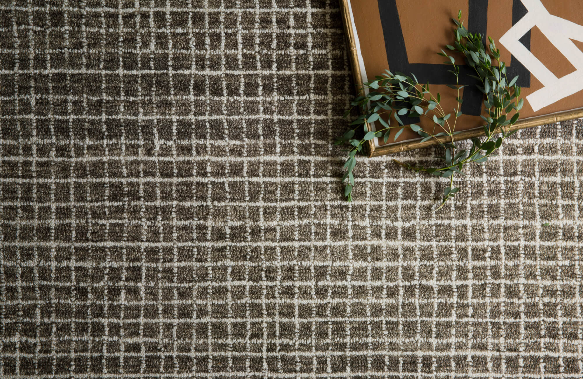Overhead detail of a graphite wool rug with a textured grid pattern, styled with framed artwork and eucalyptus branches.