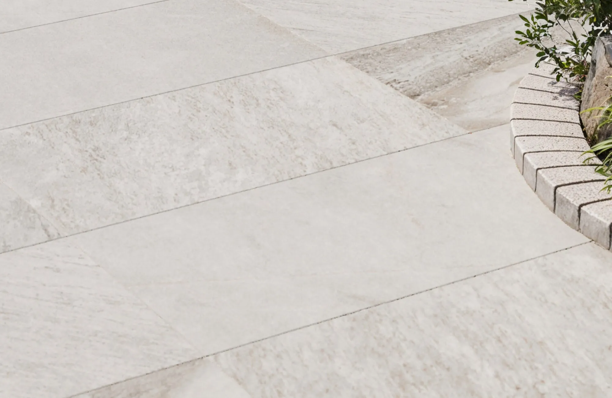 Elegant light beige porcelain outdoor paver tiles with subtle stone-like texture, arranged beside a curved garden border with greenery.