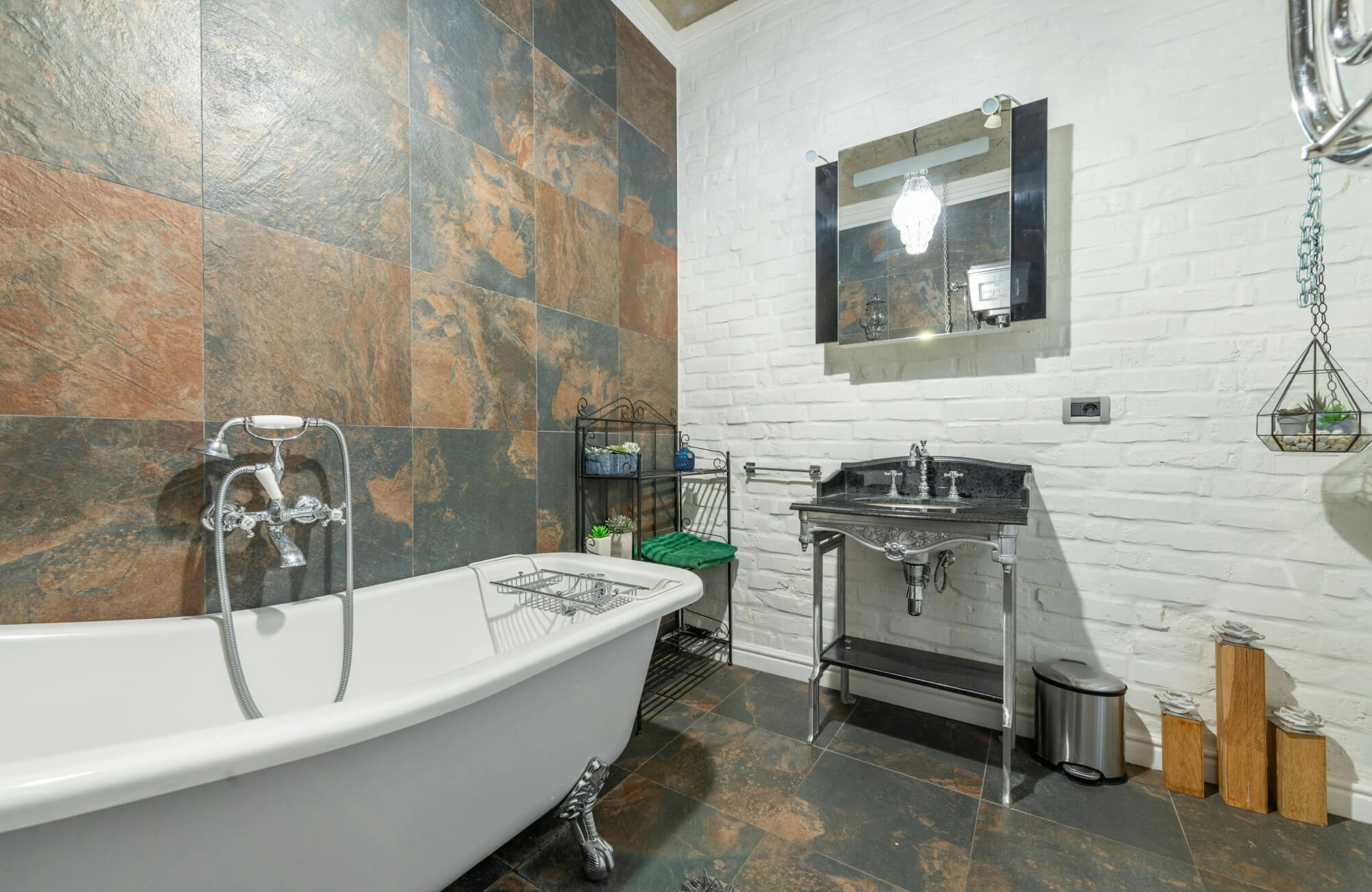 A spa-inspired bathroom with natural slate tiles in dark gray and earthy brown tones, complemented by a white freestanding bathtub, metal fixtures, and minimalist shelving.