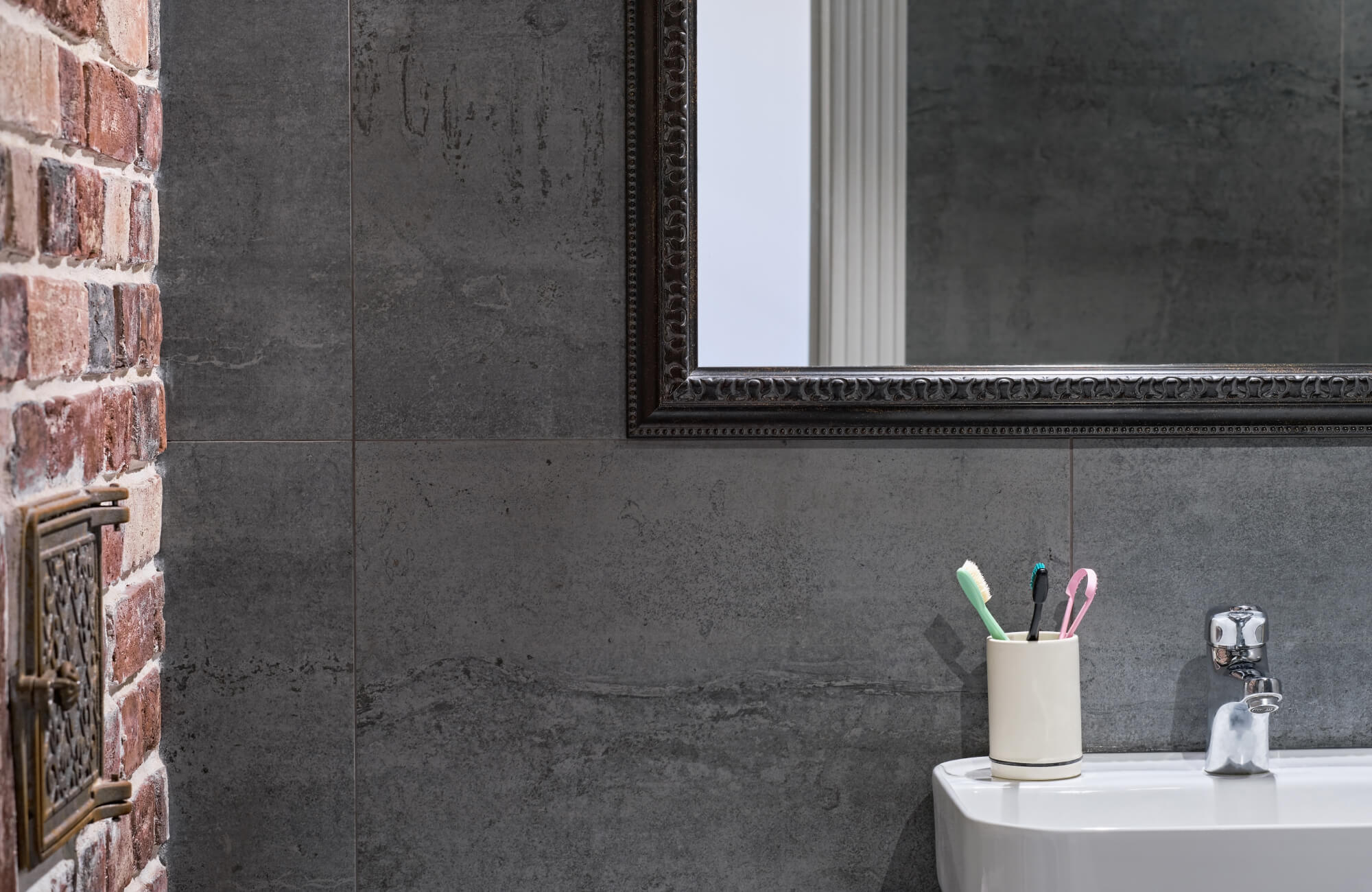 Dark gray slate wall tiles frame a compact sink area, paired with exposed brick, a vintage-style mirror, and simple bathroom accessories for a balanced, textured look.
