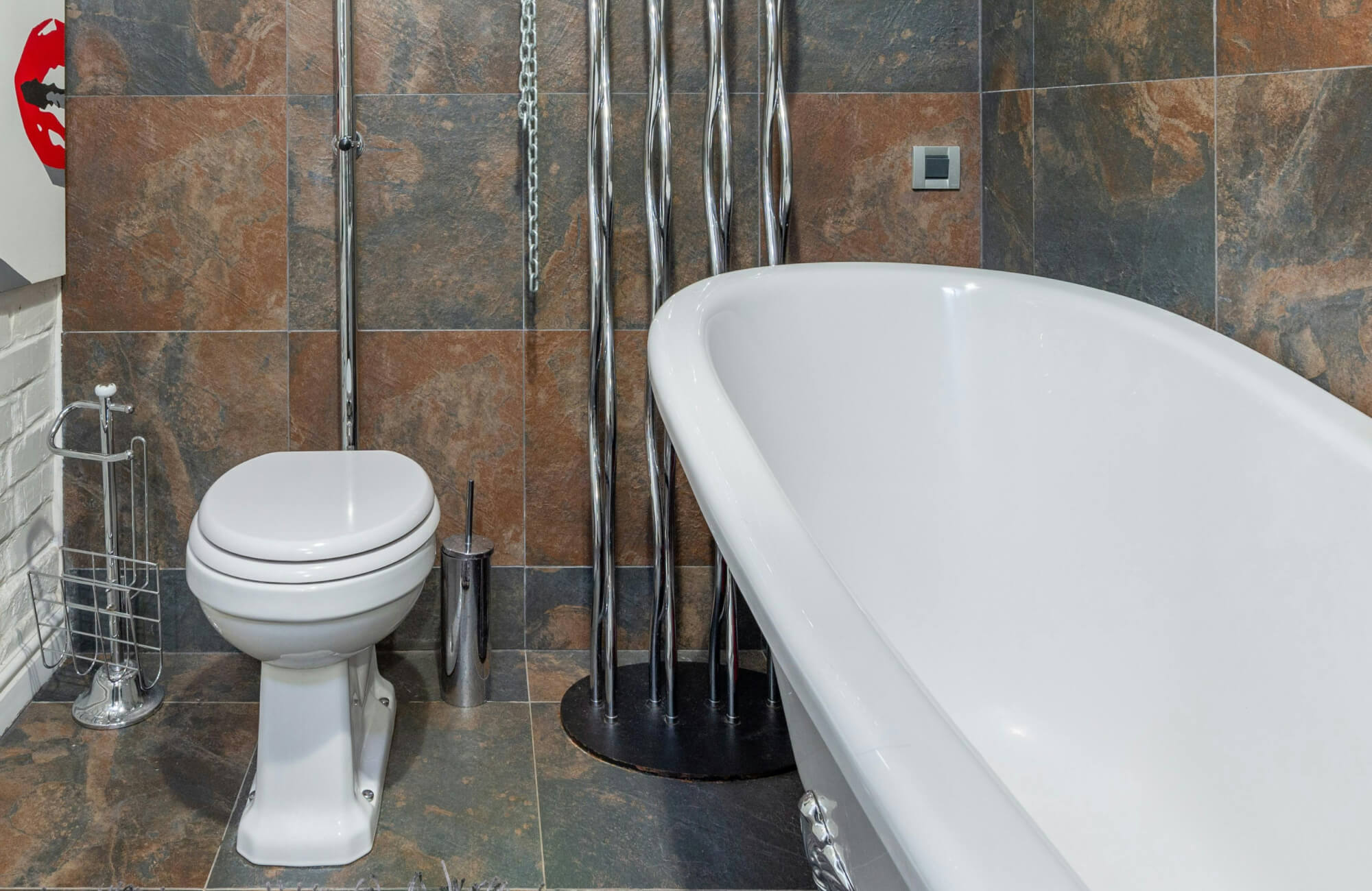 A freestanding white bathtub and toilet sit against brown and charcoal slate wall tiles, showing how natural stone surfaces hold up visually and practically in a bathroom setting.