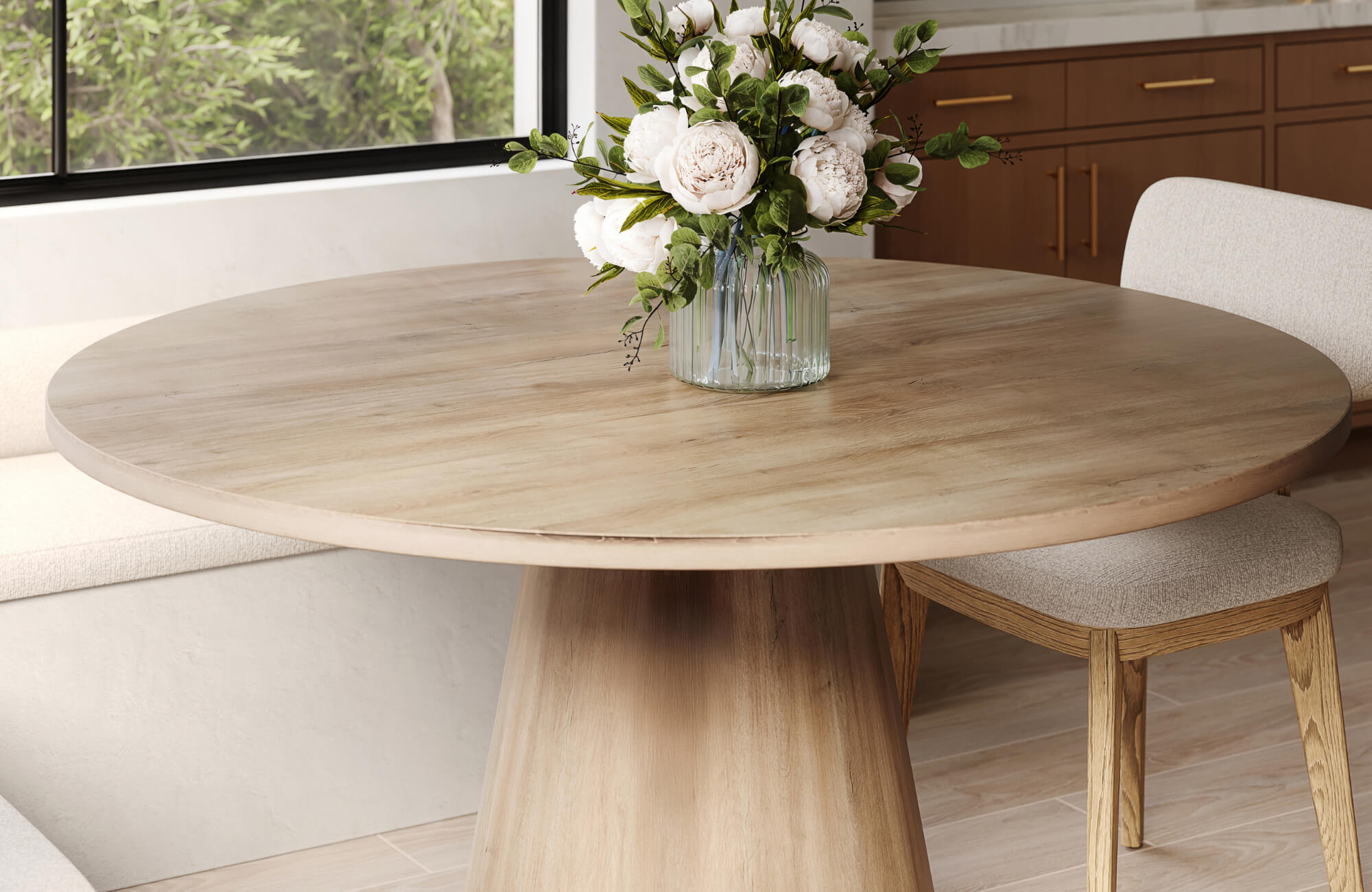 Light oak round dining table with pedestal base, styled with neutral chairs and floral centerpiece in a modern kitchen nook.