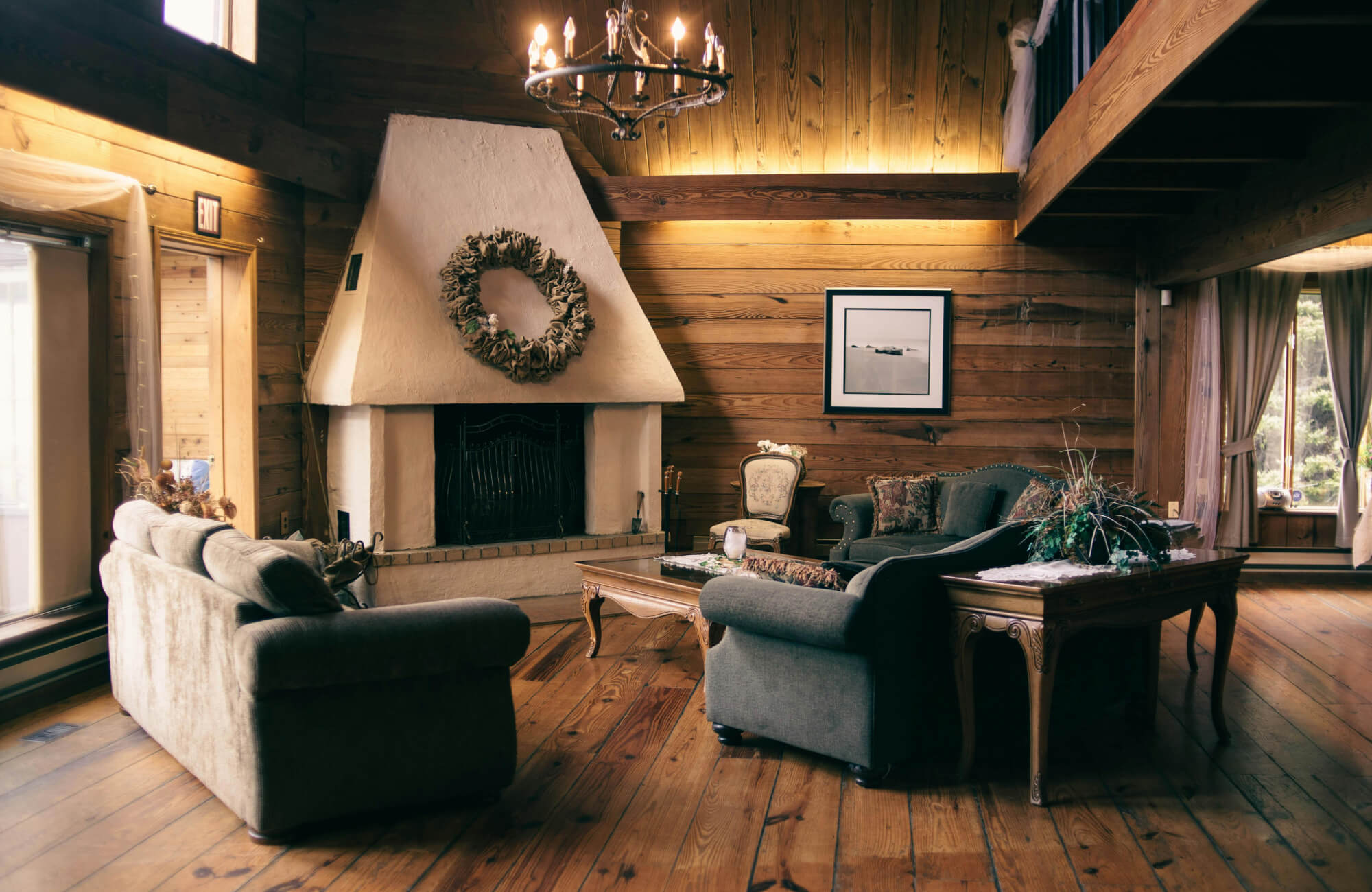 Rustic living room with natural hardwood flooring, wood-paneled walls, exposed beams, a large fireplace, vintage seating, and a chandelier.