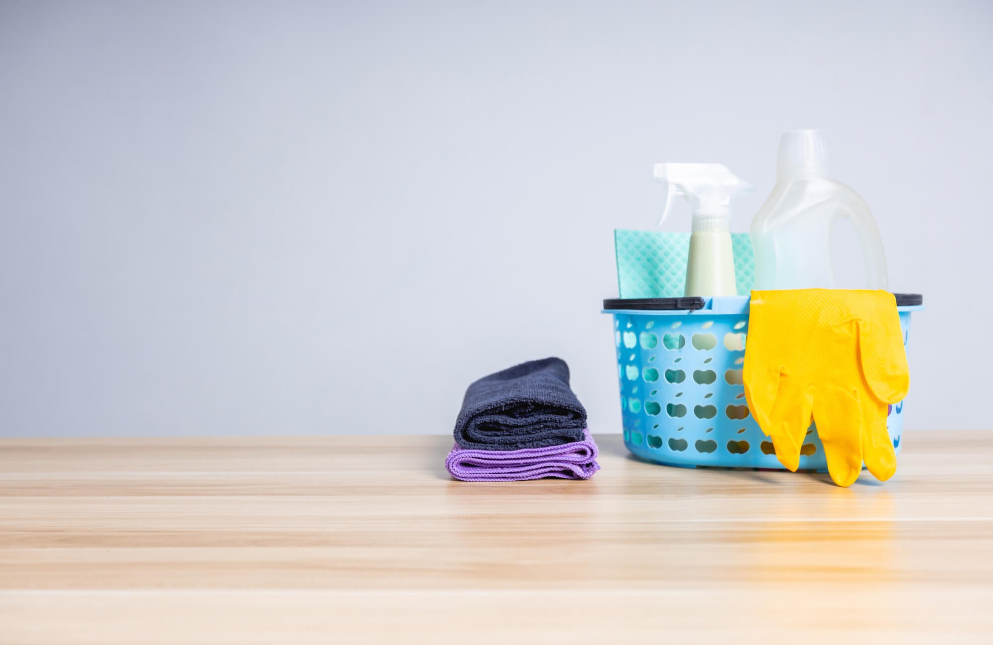 Cleaning supplies in a blue basket with yellow gloves, spray bottle, and folded towels on a light wood surface against a neutral background.
