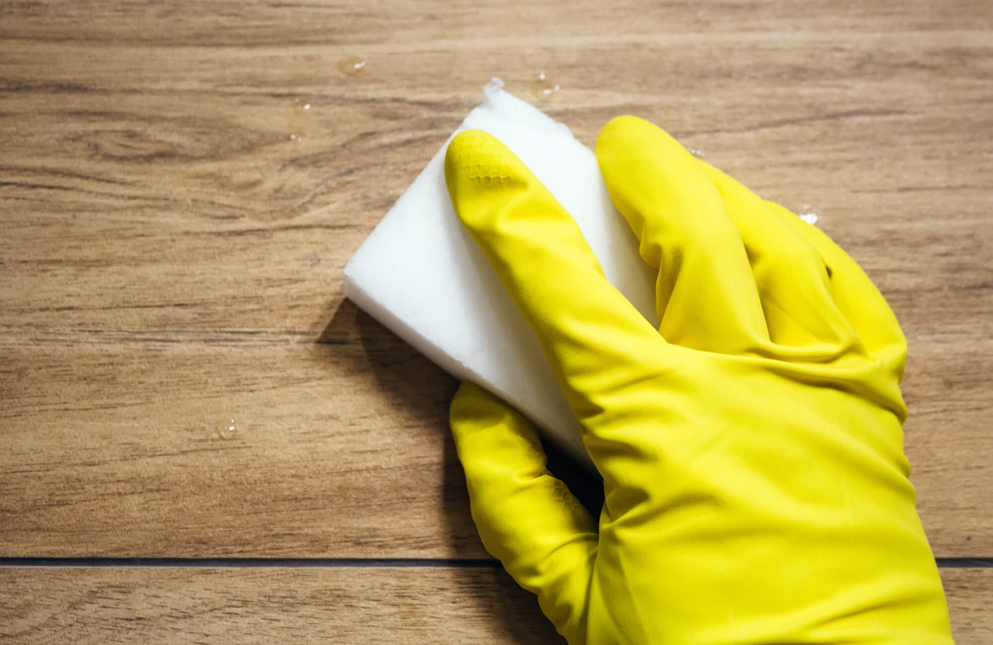 Hand wearing a yellow glove cleaning wood look tile flooring with a white sponge, highlighting easy maintenance and water resistance.