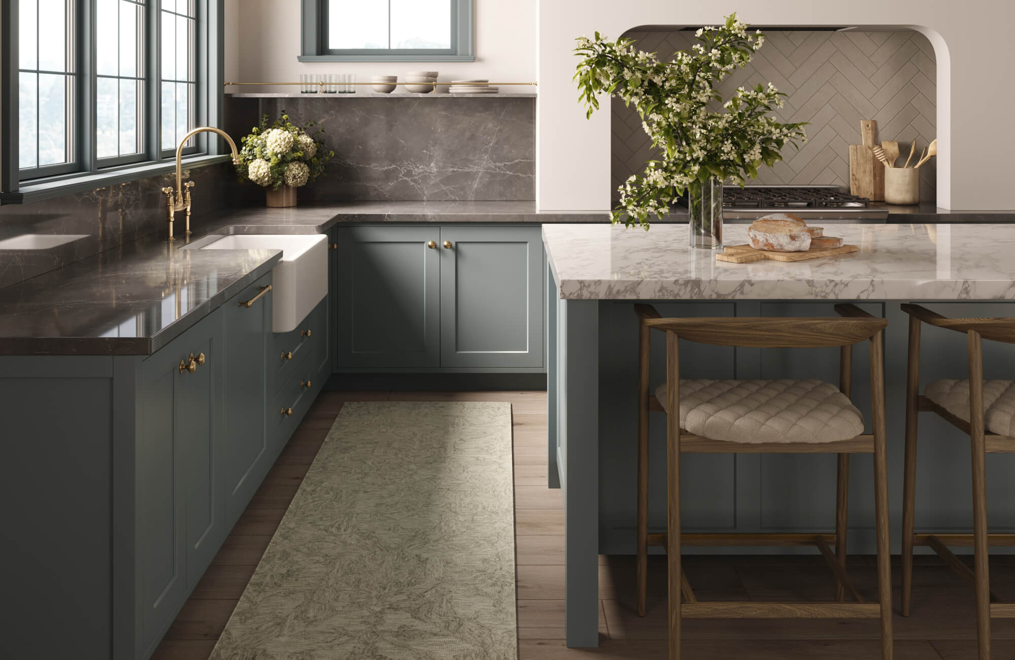 Sophisticated kitchen showcasing muted blue-gray cabinetry with brass hardware, marble countertops, a farmhouse sink with a gold-tone faucet, a herringbone tile backsplash, and a central island accented with natural wood stools and fresh greenery.
