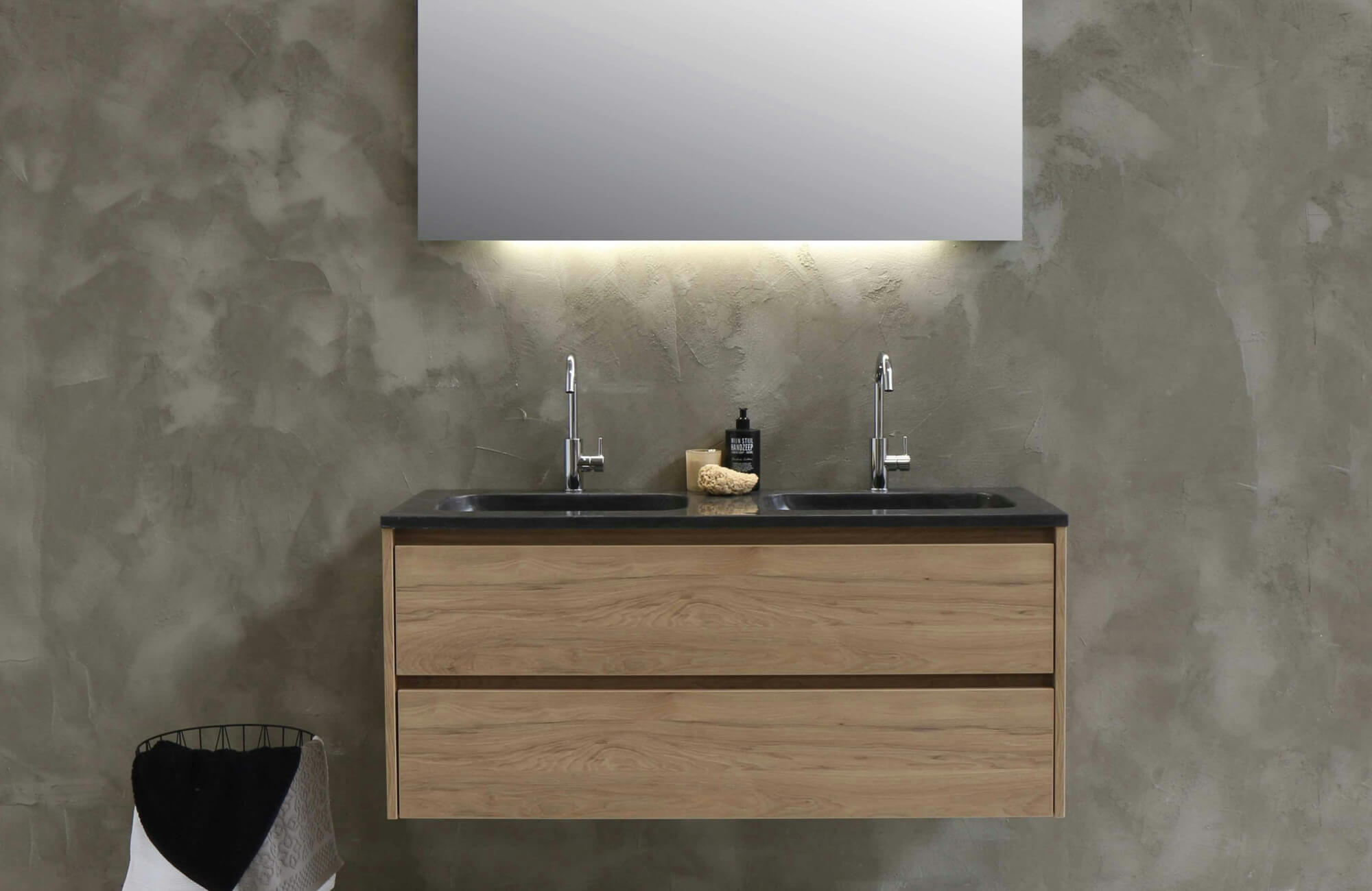 A modern bathroom vanity with floating light wood drawers and a sleek black double-sink countertop stands against a textured concrete wall, accented by minimalist chrome faucets and soft under-mirror lighting.