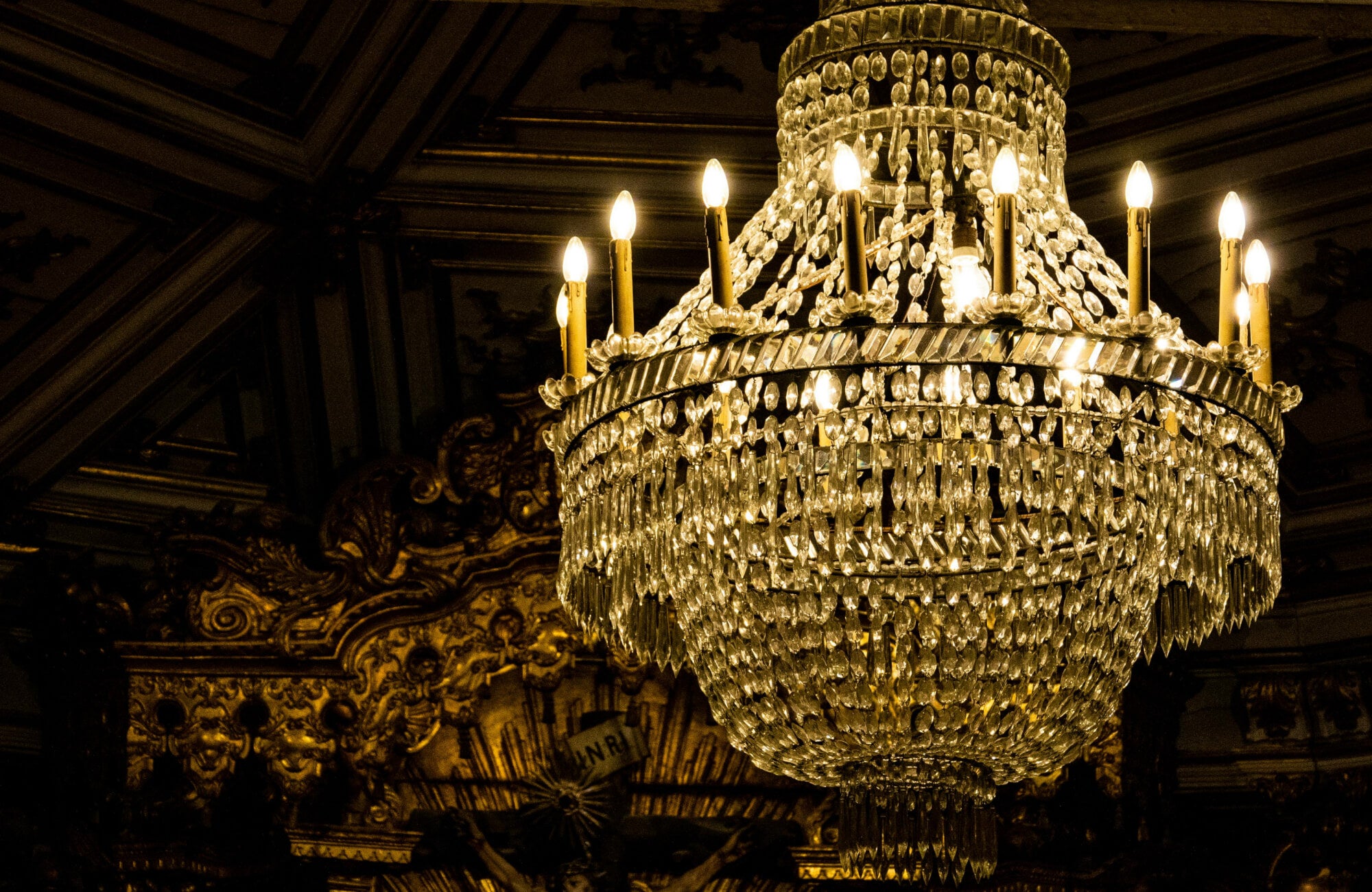 Traditional crystal chandelier adorned with cascading glass prisms and candle-style lights, radiating a warm golden glow against an ornate, gilded ceiling backdrop.