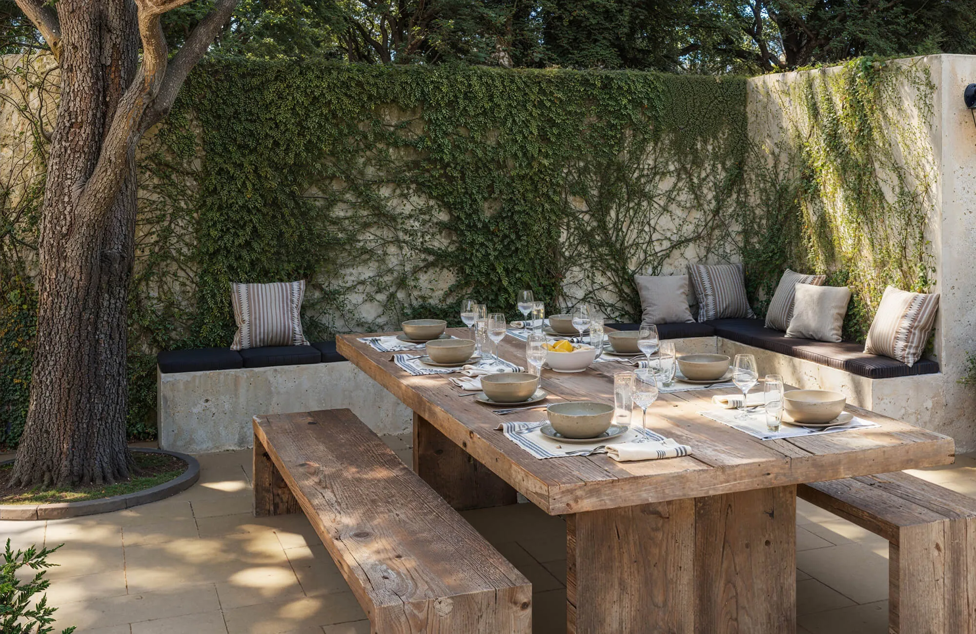 A rustic outdoor dining area features a large weathered wood table with matching benches, surrounded by built-in seating and lush ivy-covered walls beneath mature shade trees.