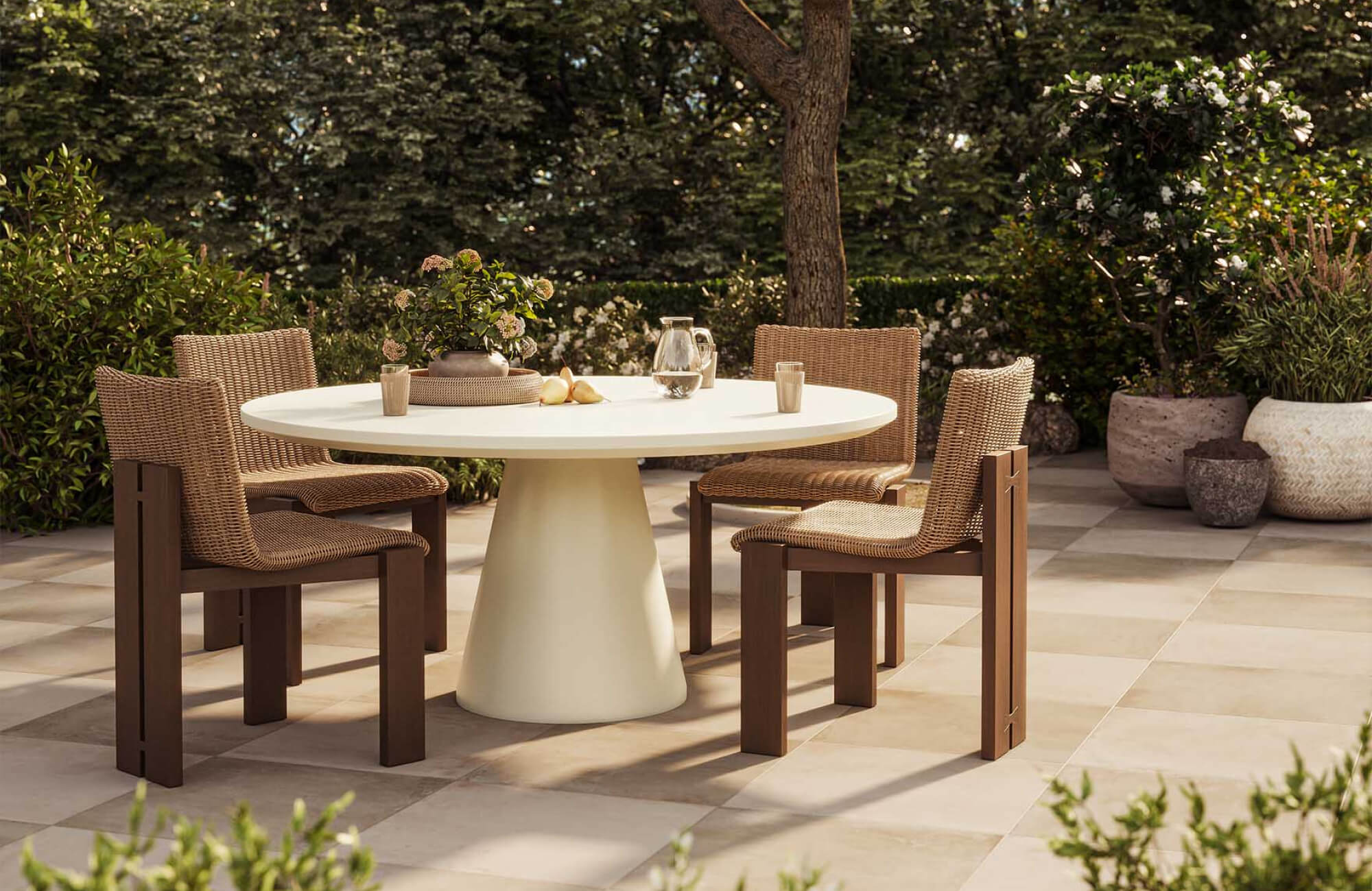 null A round pedestal dining table is positioned within a landscaped garden courtyard, surrounded by woven dining chairs and flowering greenery.