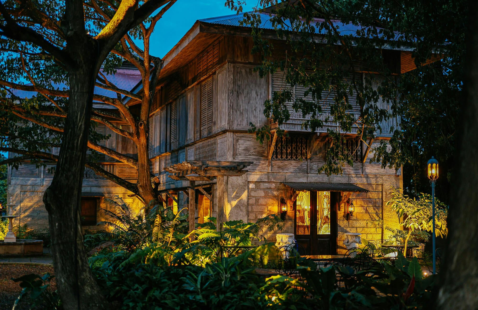Atmospheric outdoor house lighting with warm wall-mounted lanterns and garden path lights illuminates this rustic home, highlighting weathered wood textures, stone details, and surrounding greenery at dusk.