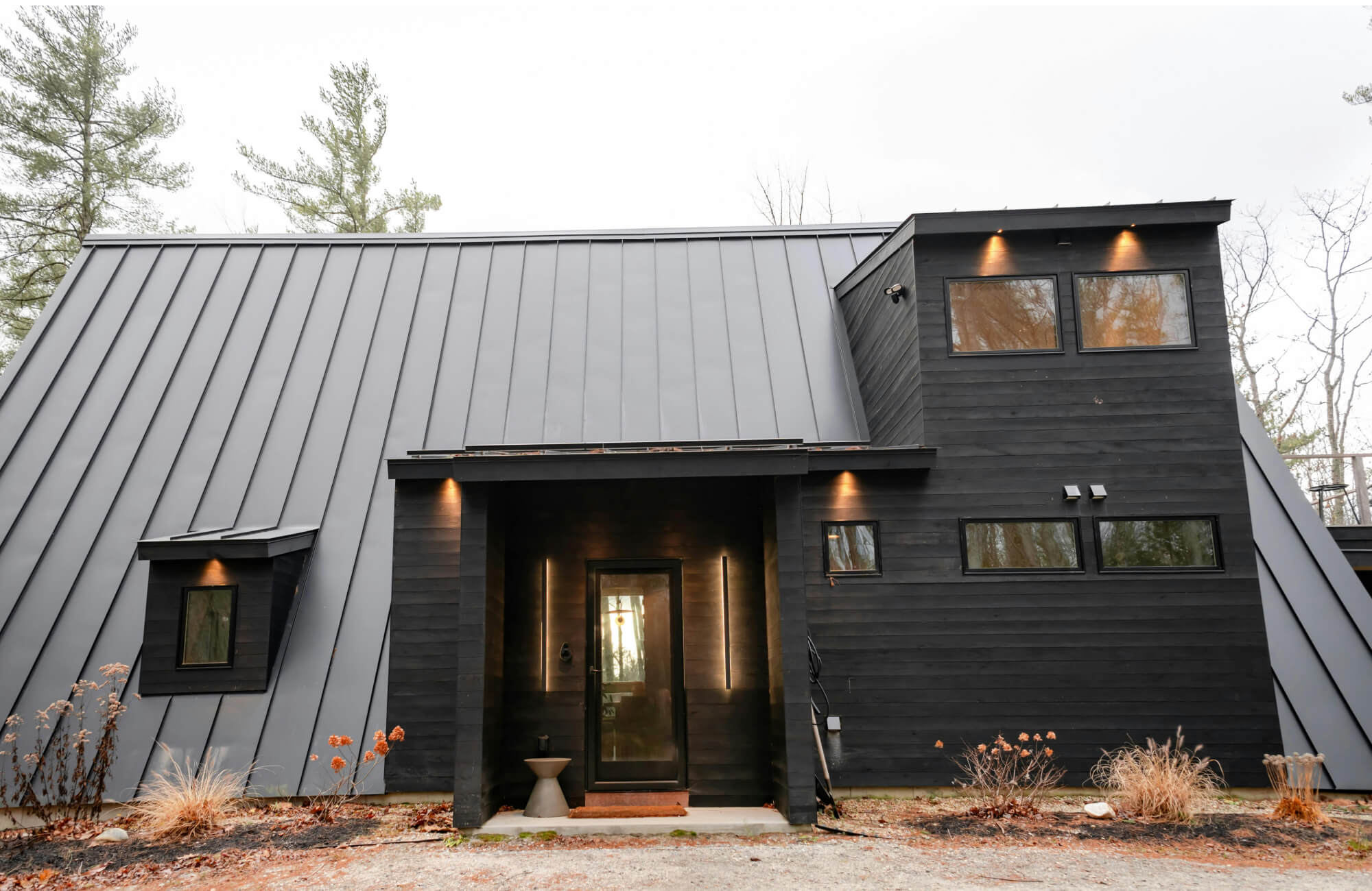 Minimalist outdoor house lighting with recessed downlights and vertical LED entry lights accentuates this modern black-clad home, emphasizing the angular roofline, clean façade, and inviting front entrance.