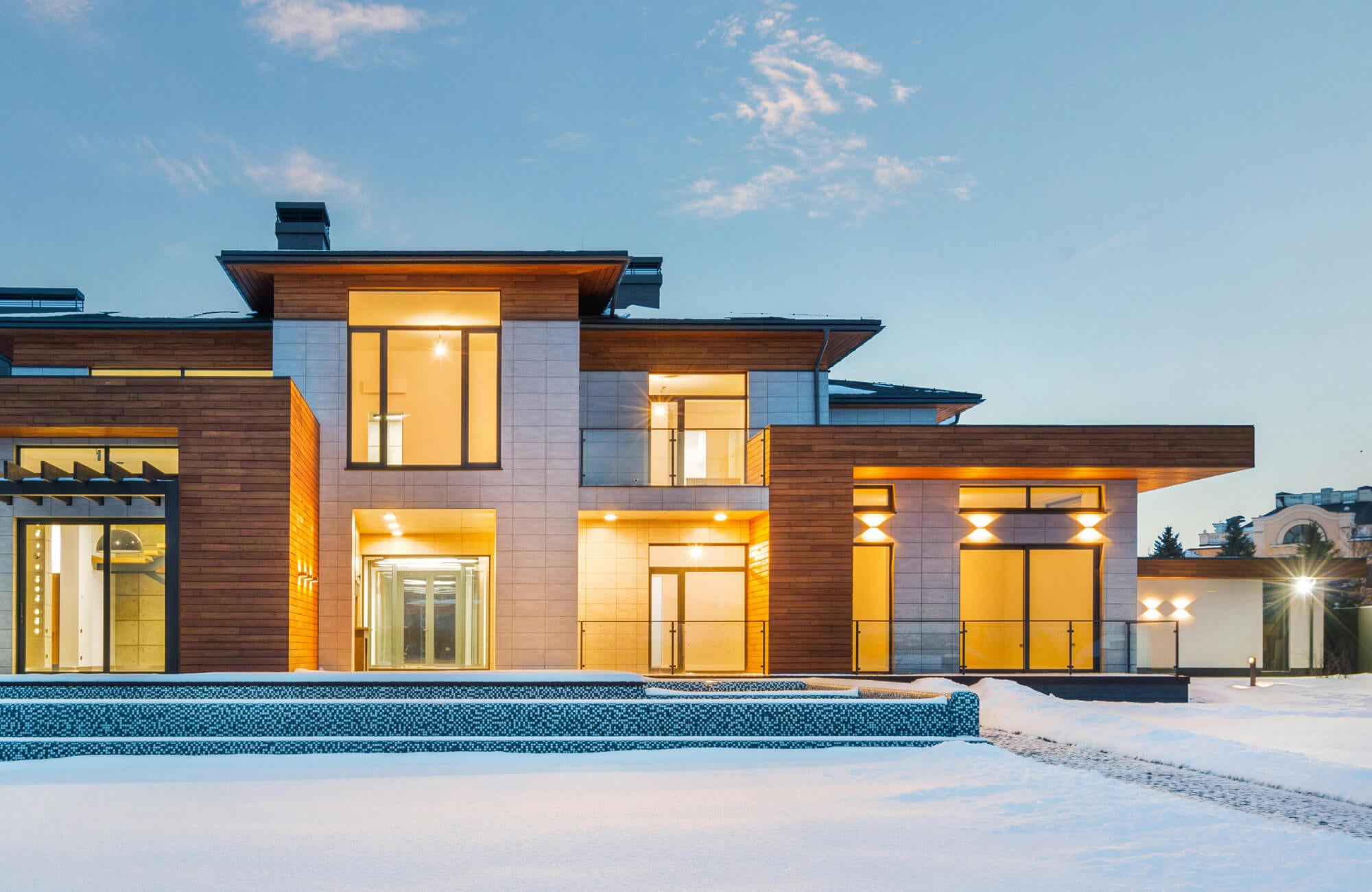 Contemporary outdoor house lighting with wall-mounted sconces and recessed soffit lights bathes this modern home in warm illumination, highlighting layered volumes, wood accents, and expansive glass against the winter landscape.