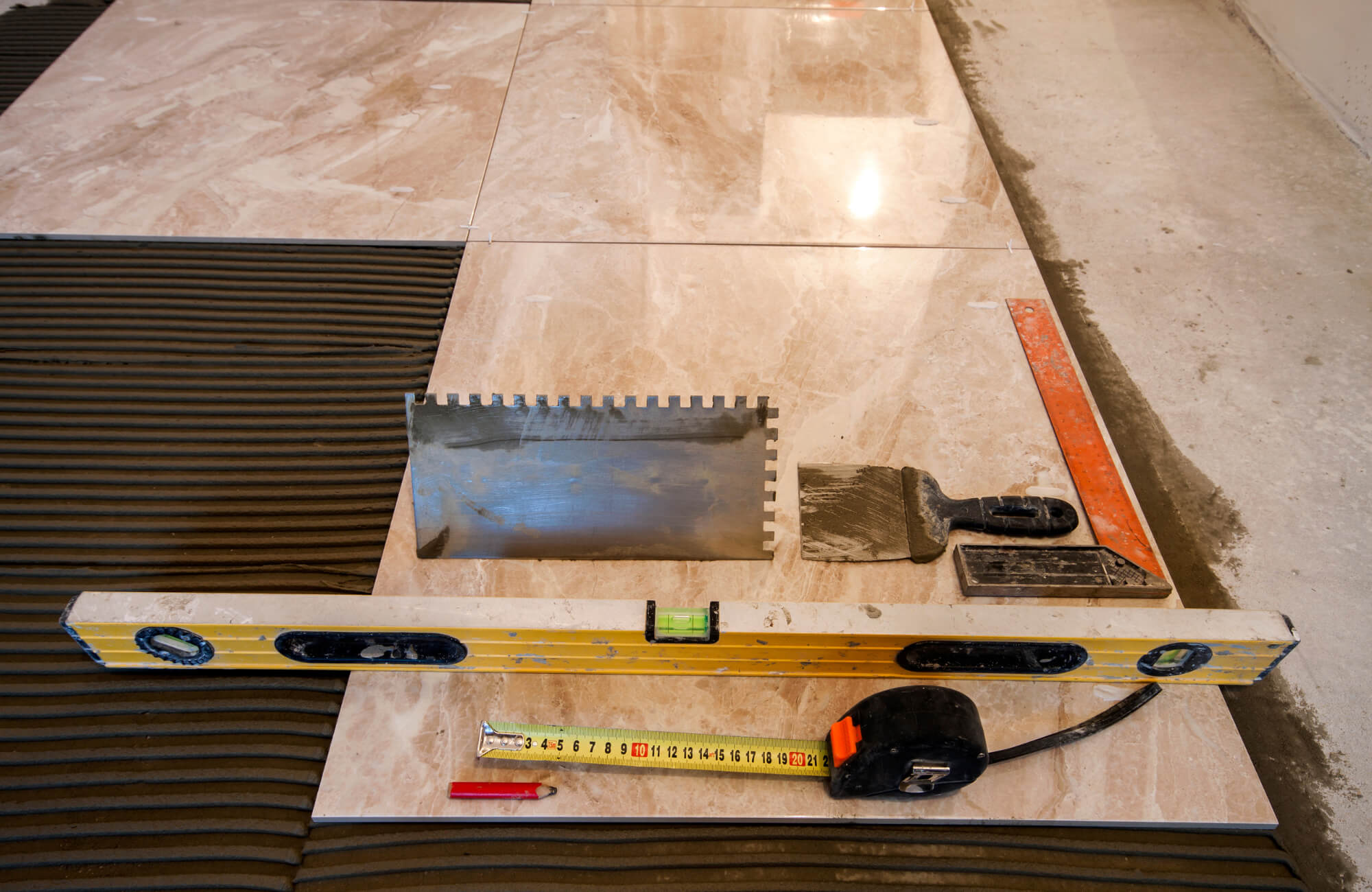 null Tile installation in progress with beige marble-look tiles, notched trowel, level, measuring tape, and other tools on a mortar bed.