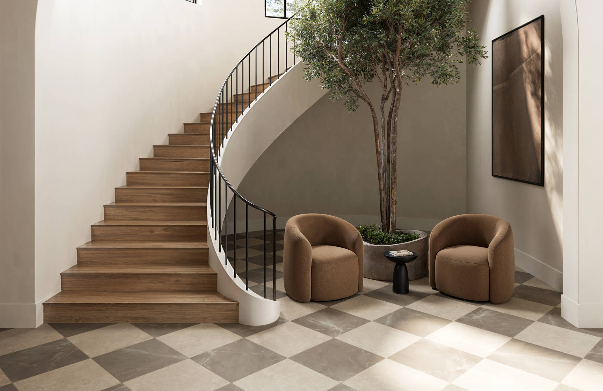 Grand staircase entryway with warm wood steps, taupe checkered floor tiles, and cozy lounge chairs framing an indoor tree.