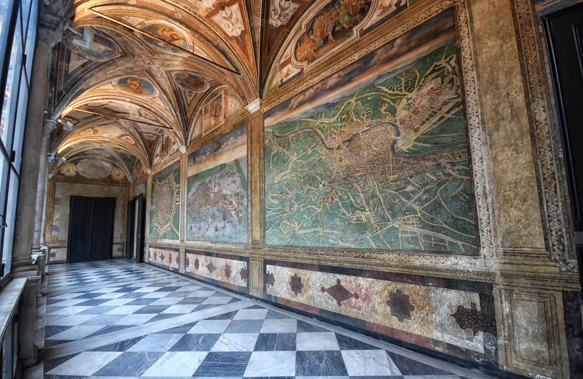 Historic gallery with vaulted frescoed ceilings, large map murals, and a diagonal black-and-white marble checkerboard tile floor.