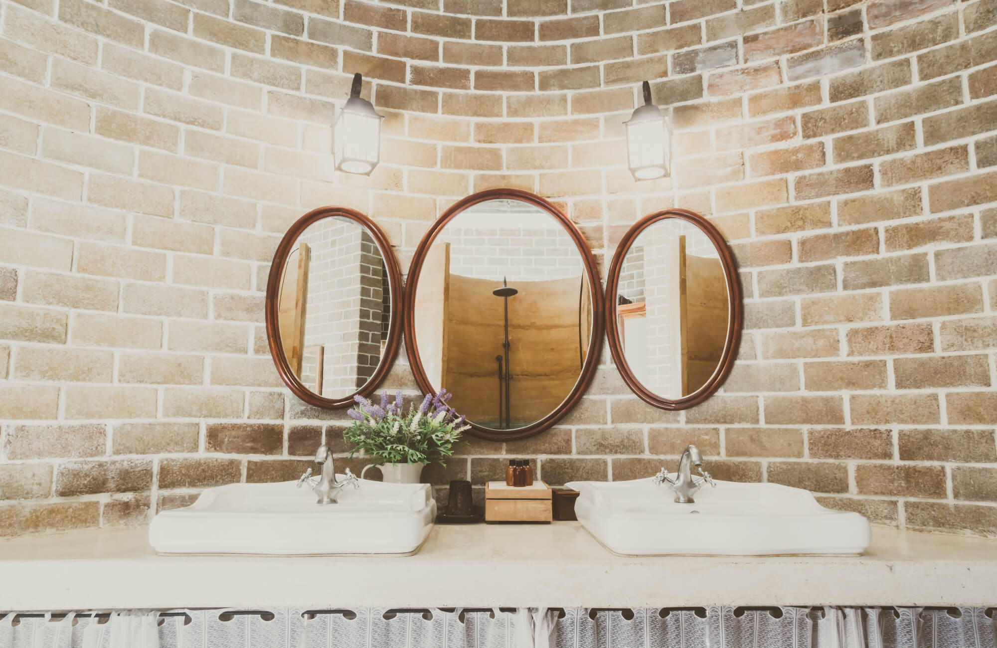 null Round wooden-framed oval mirrors mounted against a curved brick wall above a double sink vanity, with lantern-style sconces adding a warm, rustic touch.