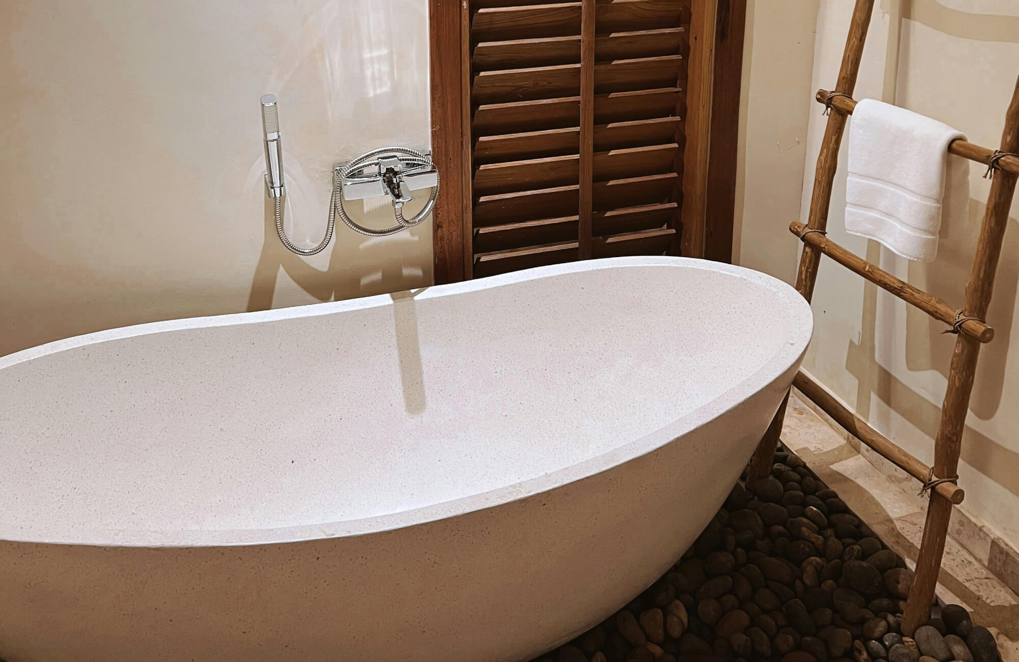 Minimal rustic bathroom with a stone-look freestanding tub, wood towel ladder, and pebble flooring accents.