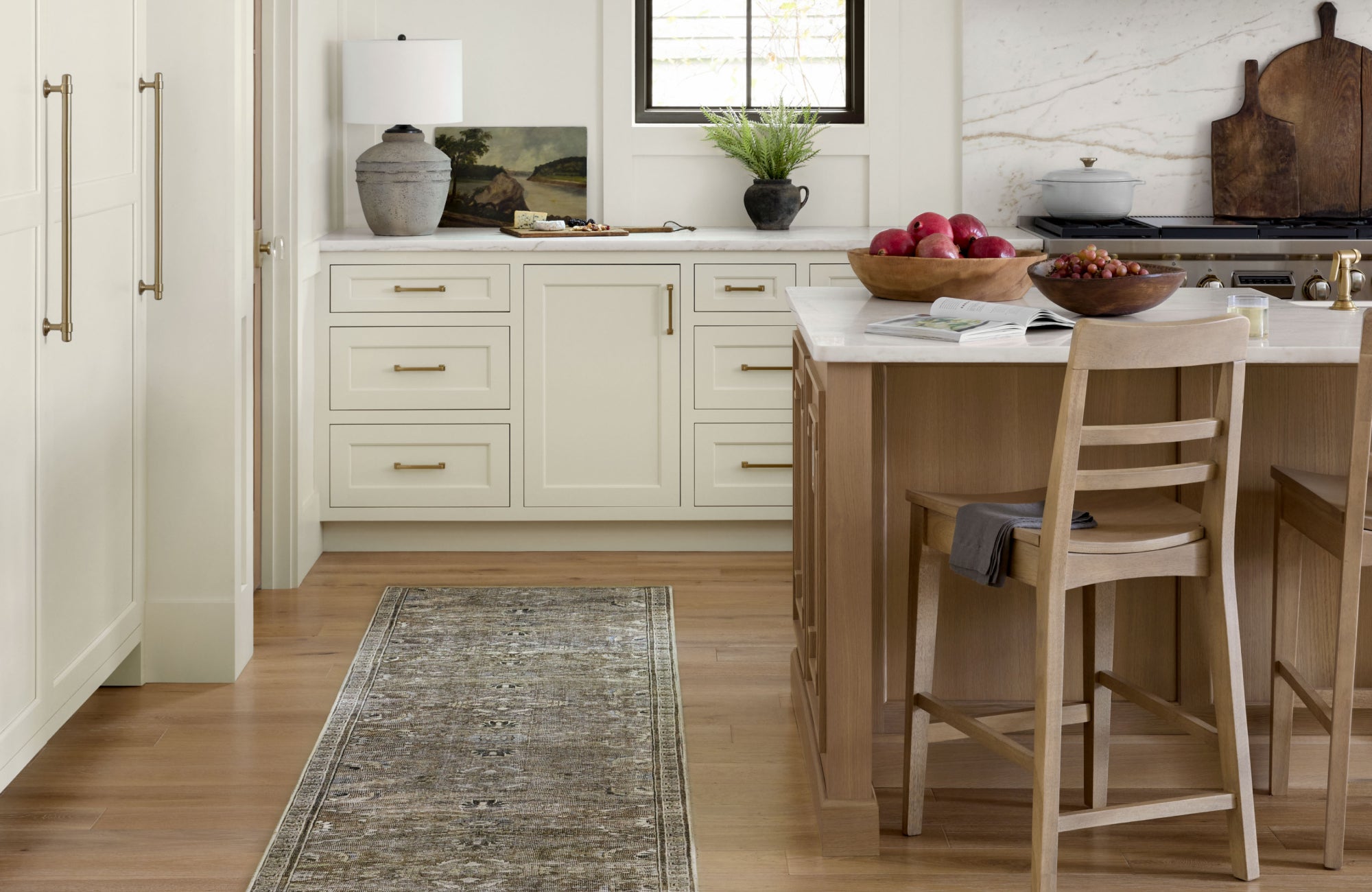 A soft, neutral kitchen featuring white cabinetry, wood accents, and a dark-toned runner rug that grounds the space with contrast and vintage charm.