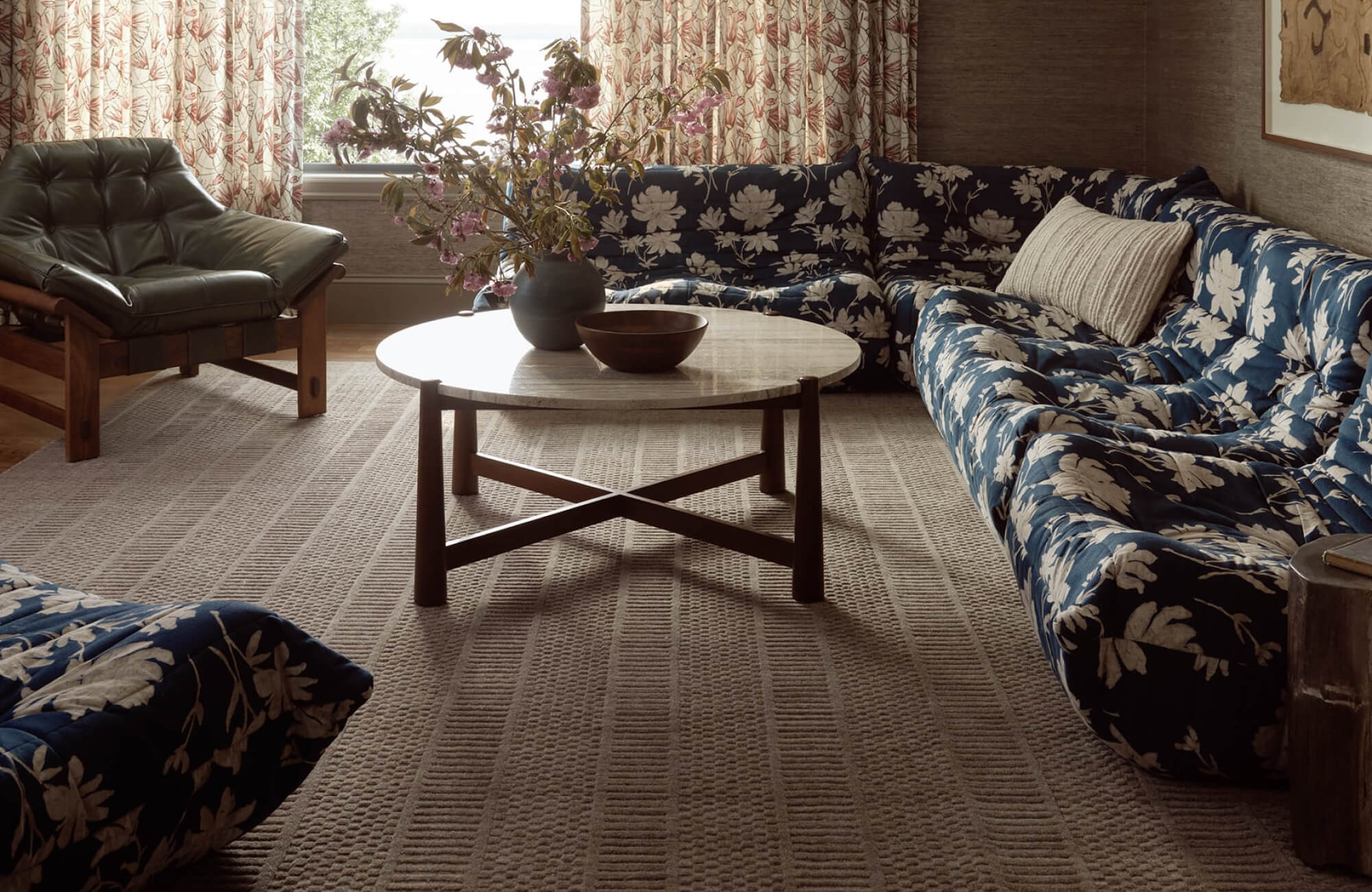 Cozy living room with a deep blue floral sectional, tufted leather lounge chair, and textured neutral rug.