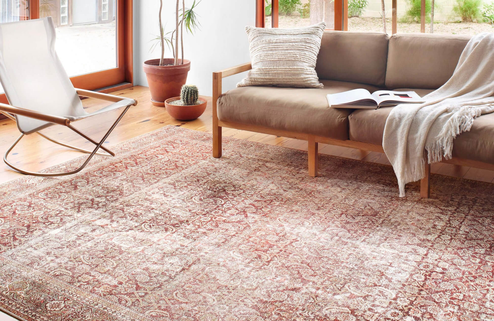 Sunlit living space with a distressed red vintage rug, wood-frame sofa, potted plants, and warm-toned plank flooring.