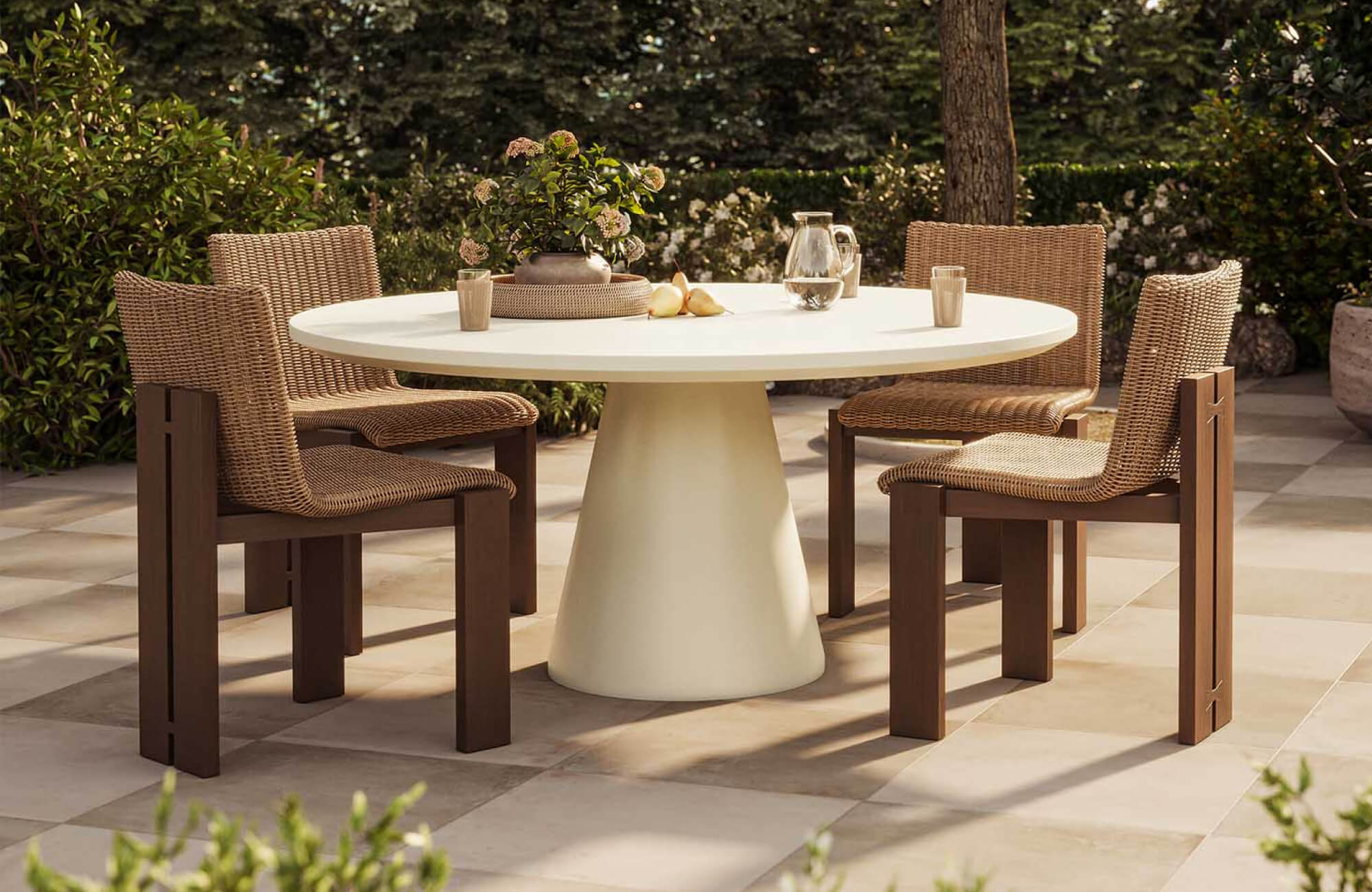 Warm beige and white outdoor floor tiles surround a round dining table with woven chairs in a lush garden patio.