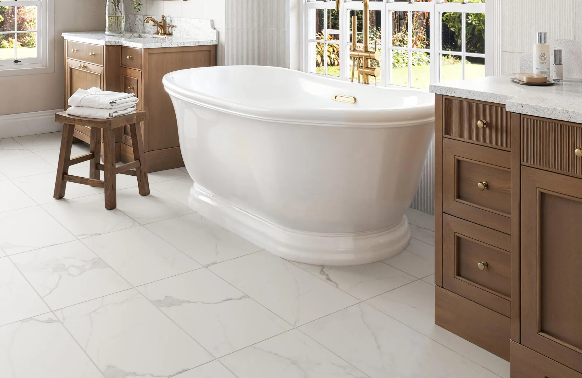 Light marble-look bathroom floor tiles with soft gray veining beneath a freestanding tub, a wooden stool, and a warm-wood vanity create a bright, open feel that makes the space appear larger.