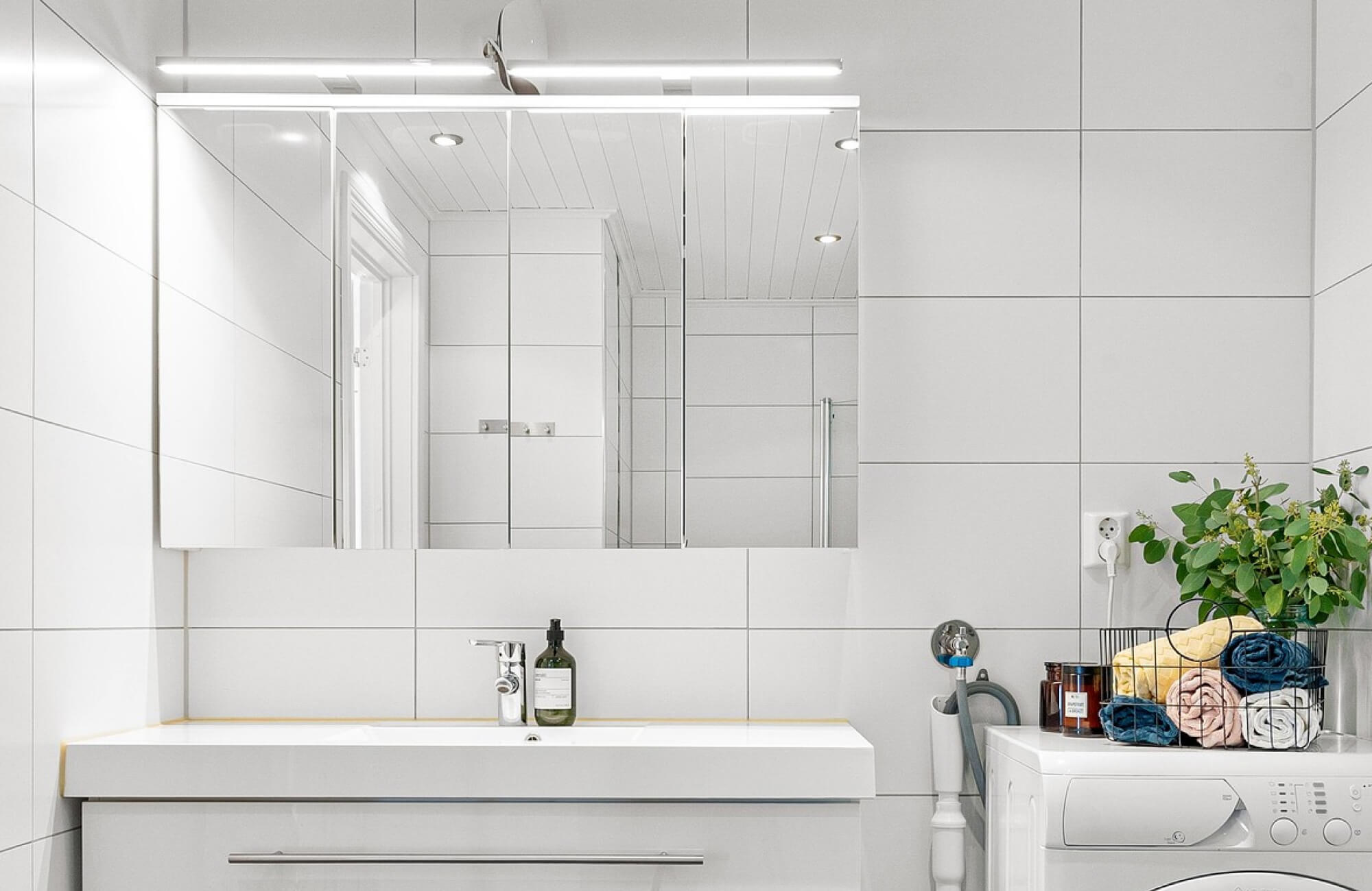 Minimalist bathroom with a sleek frameless mirror, glossy white tiles, and a modern vanity beside a laundry station.
