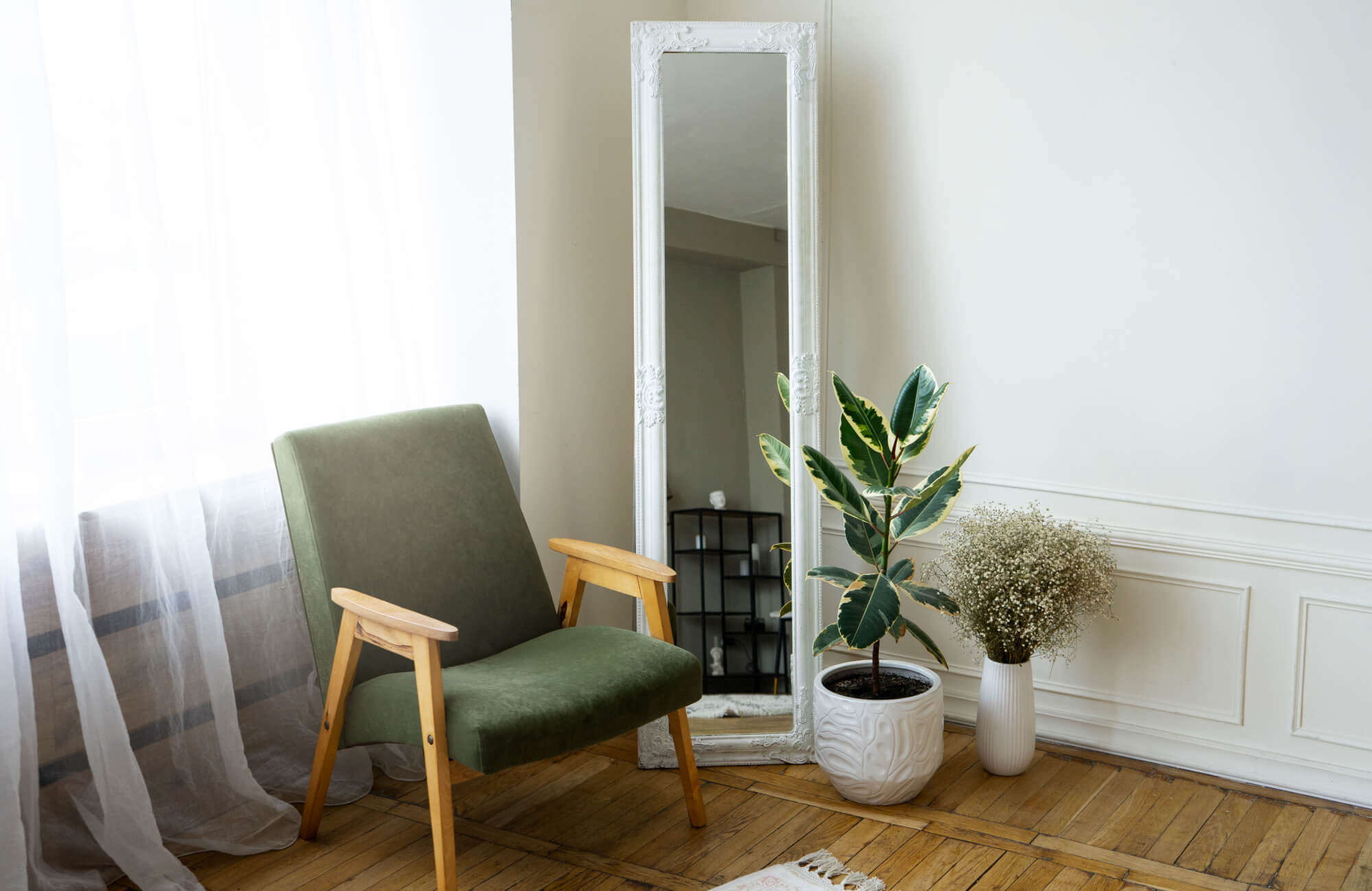 A white-framed floor mirror rests against the wall next to a green chair, with a leafy plant and delicate dried florals adding a cozy, styled touch to the room.