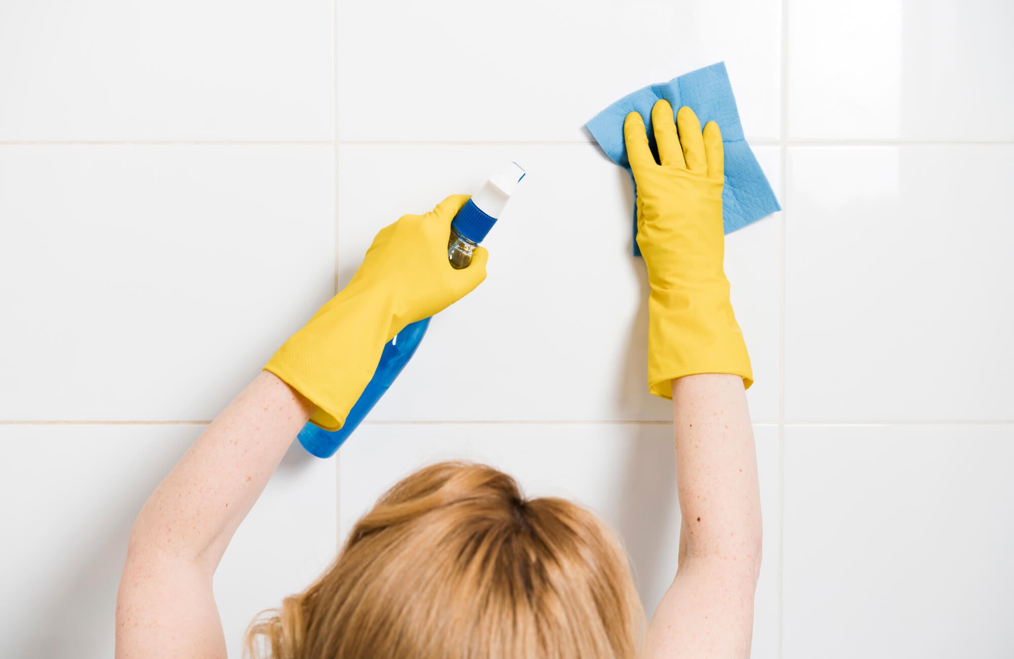 null Woman cleaning glossy white tiled bathroom walls with a spray bottle and cloth, wearing yellow rubber gloves for a sparkling, streak-free finish.