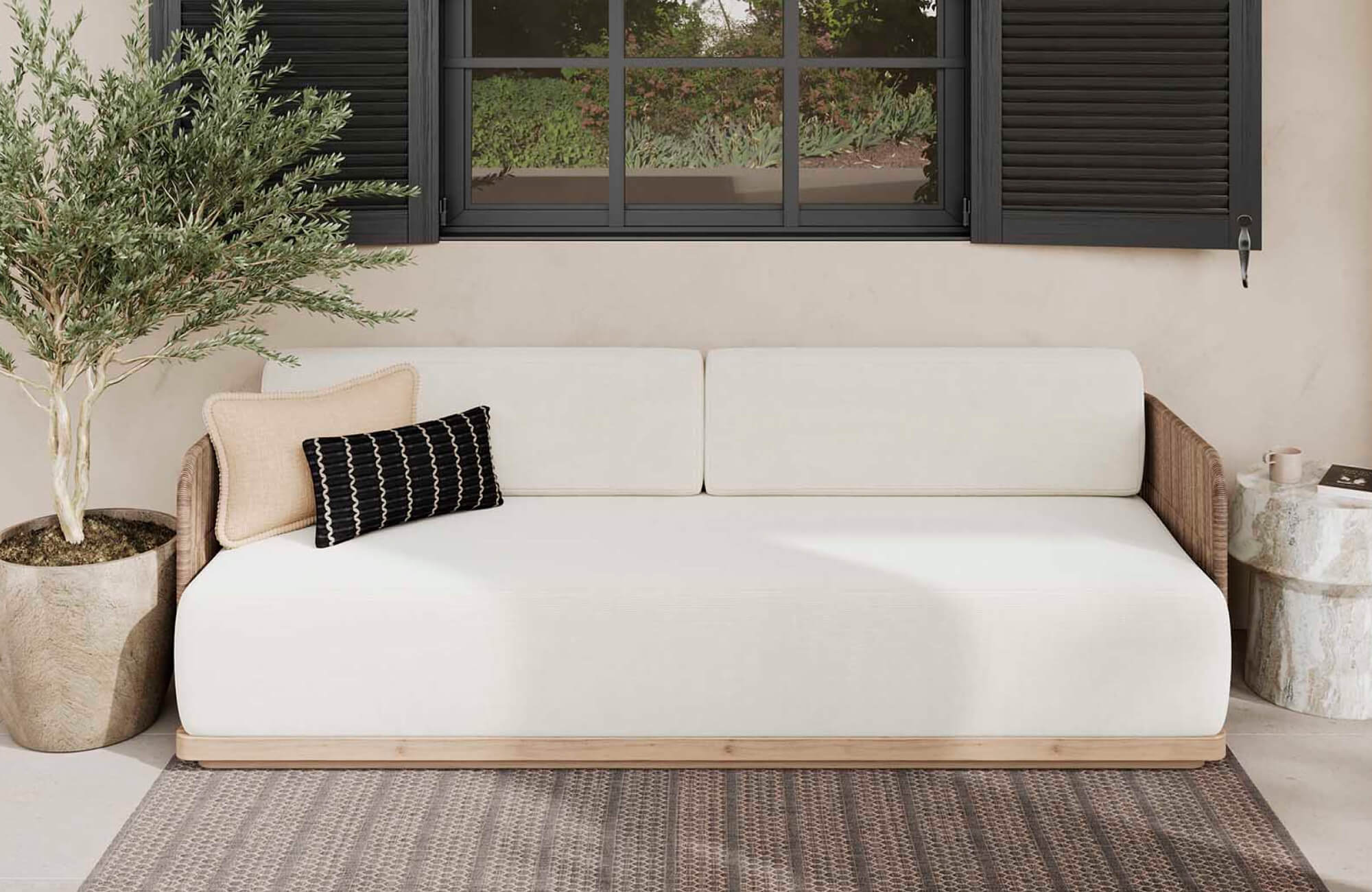 Minimal outdoor sofa with white cushions, a beige square pillow, and a black patterned lumbar pillow placed below a window with black shutters.