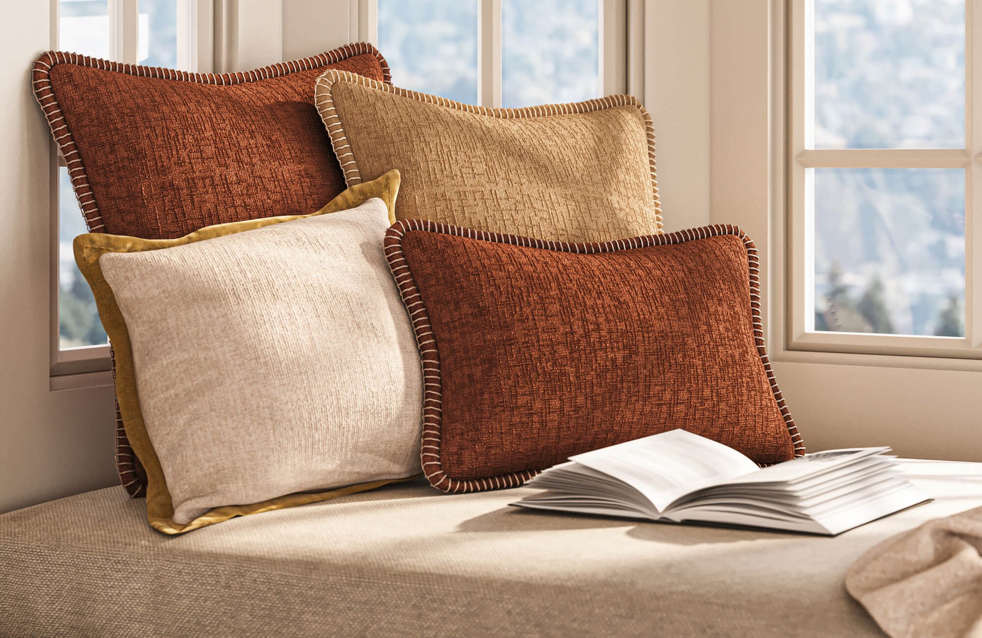Sunlit window seat styled with layered textured throw pillows in rust, tan, cream, and ochre tones beside an open book.
