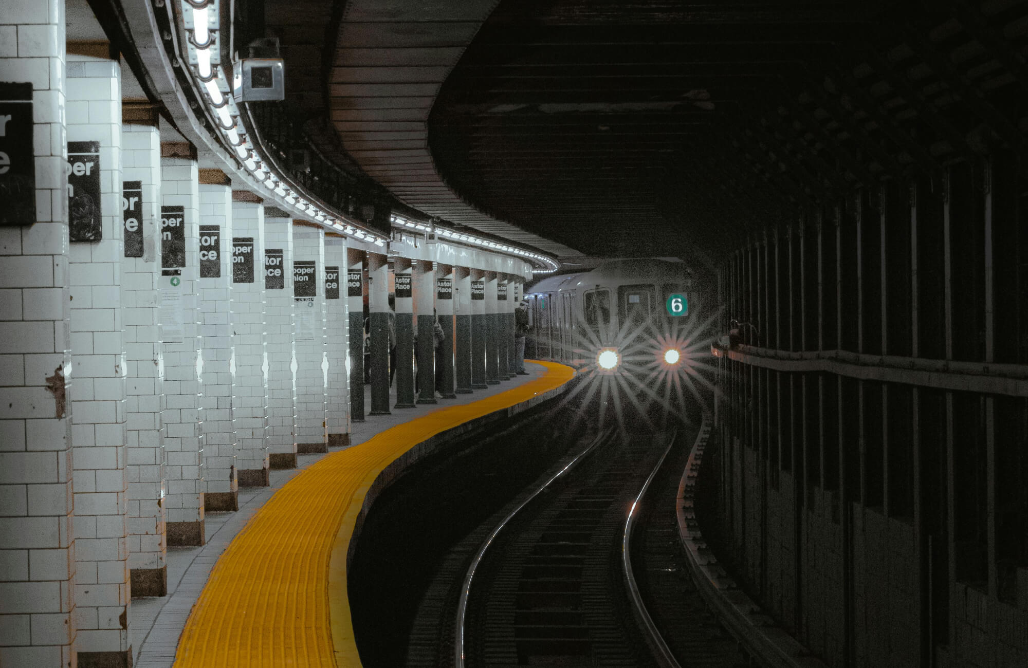 A New York subway train approaches a station with white subway tiles lining the curved walls and a bright yellow tactile paving along the platform edge