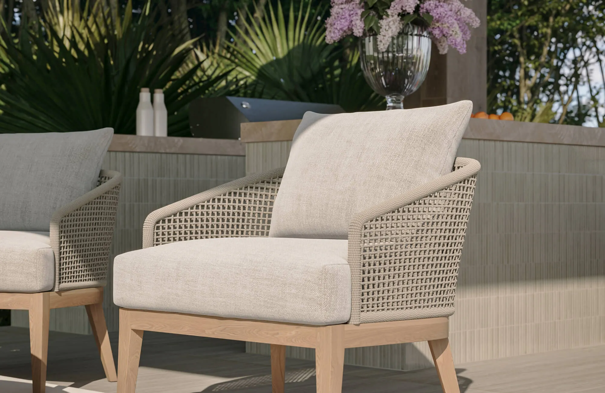 Close-up of a woven outdoor lounge chair with cream cushions beside a tiled outdoor kitchen counter surrounded by lush tropical landscaping.