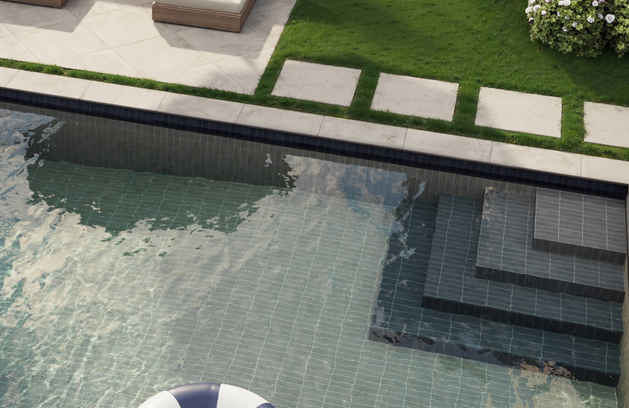 Modern pool design featuring charcoal-toned step tiles and creamy stone pavers bordered by lush green grass.