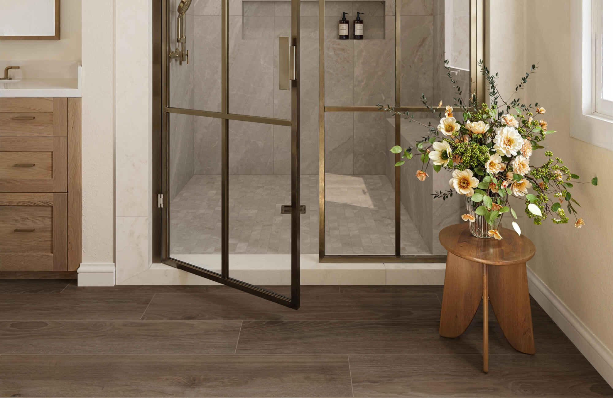 Warm bathroom with dark brown wood-look floor tiles, glass shower enclosure, brass fixtures, and soft neutral wall finishes.