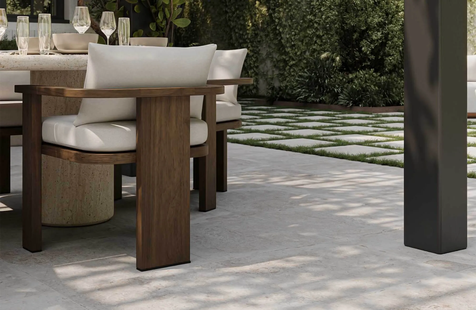 Modern outdoor dining patio with wood lounge chairs, stone table, and large format pavers framed by lush garden greenery nearby.