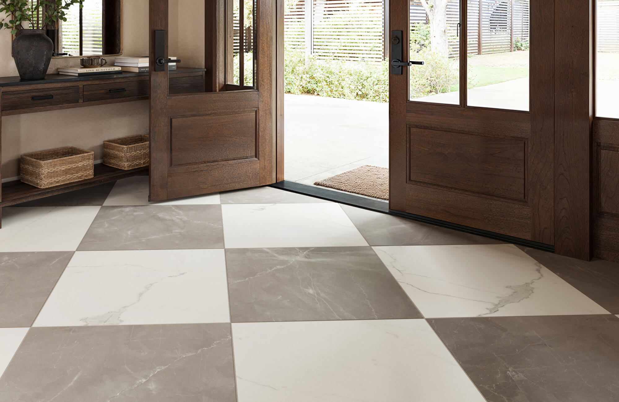 A stylish entryway featuring large checkered tiles in shades of white and grey, complemented by dark wooden doors and a console table with woven baskets.