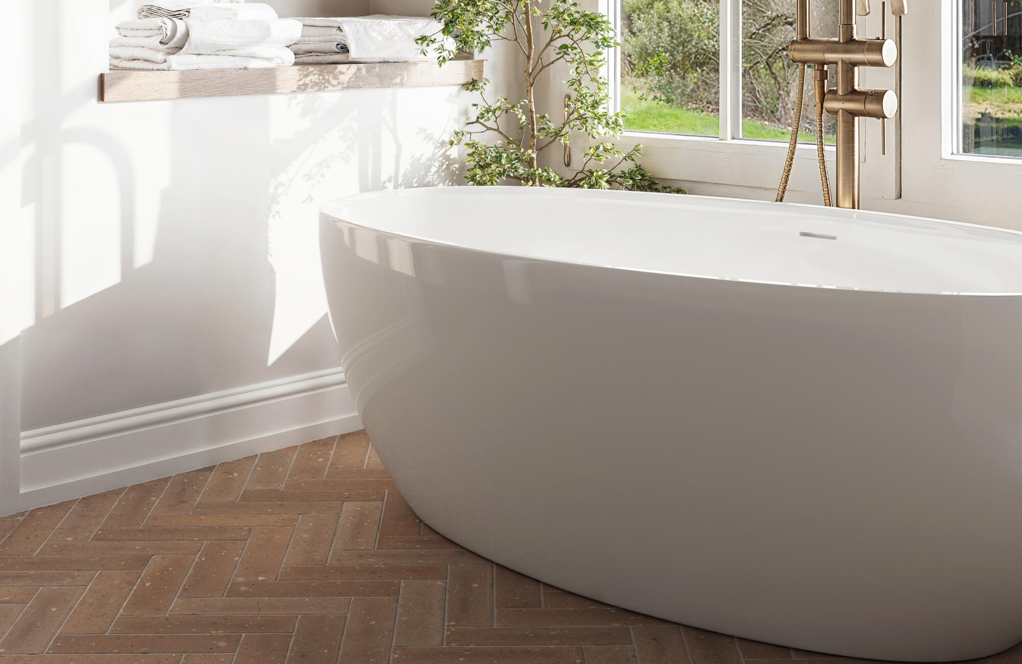 Terracotta herringbone subway tile flooring adds texture and slip resistance, complementing a sleek freestanding tub and brass fixtures in a sunlit bath.