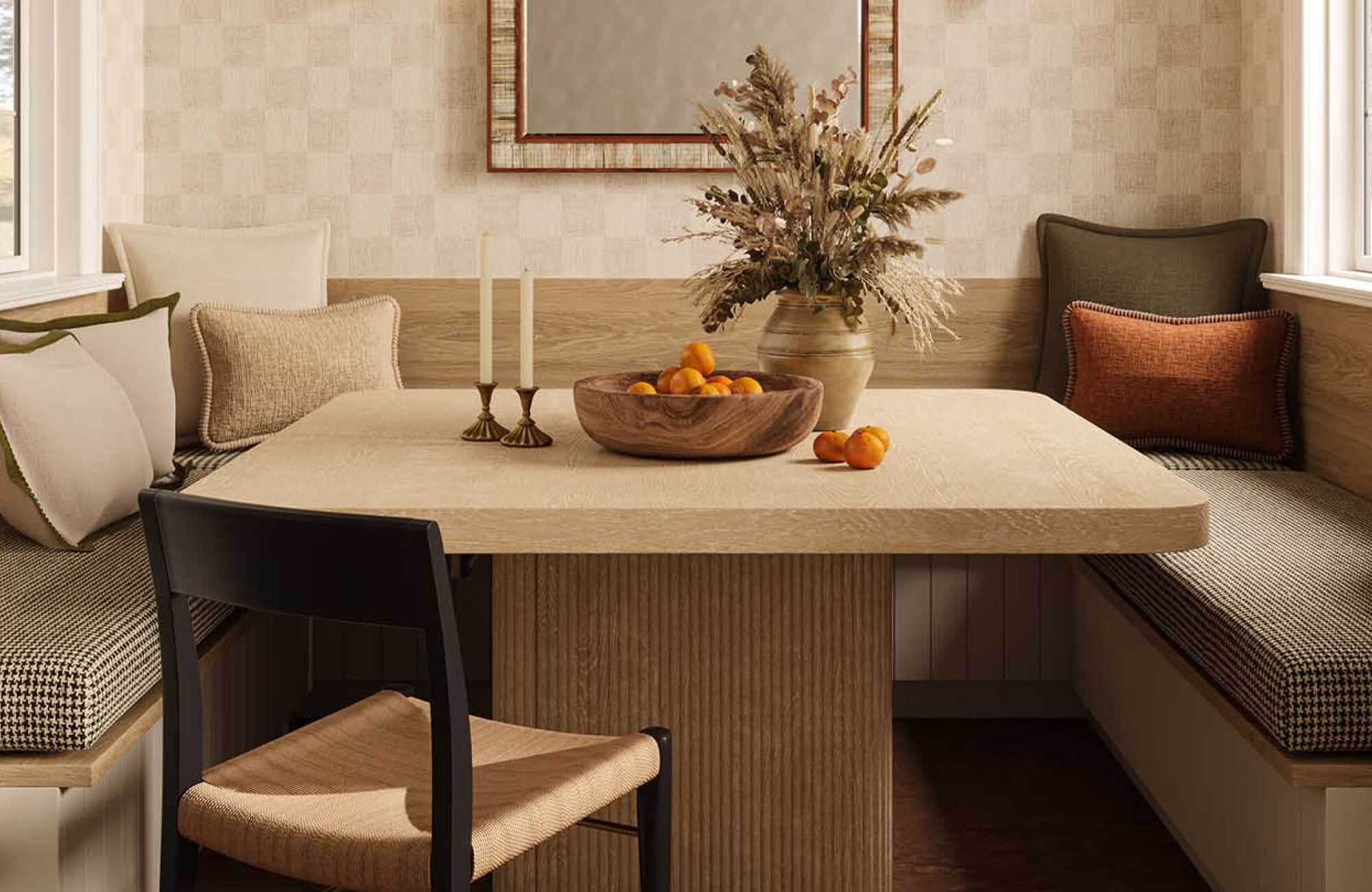 null Wooden bowl filled with oranges on a dining table in a cozy breakfast nook with banquette seating, candles, and a dried floral arrangement.